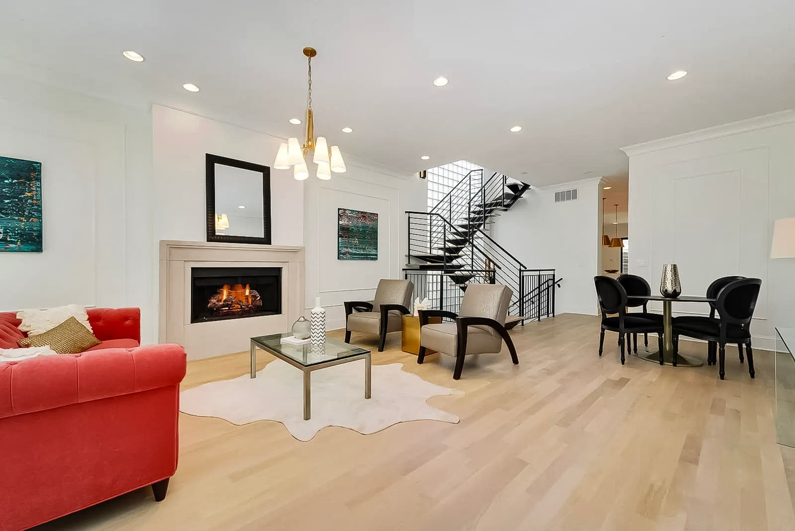 Salon moderne avec cheminée encastrée, canapé rouge élégant, fauteuils beiges, escalier en métal noir, sol en bois clair et éclairage suspendu doré.