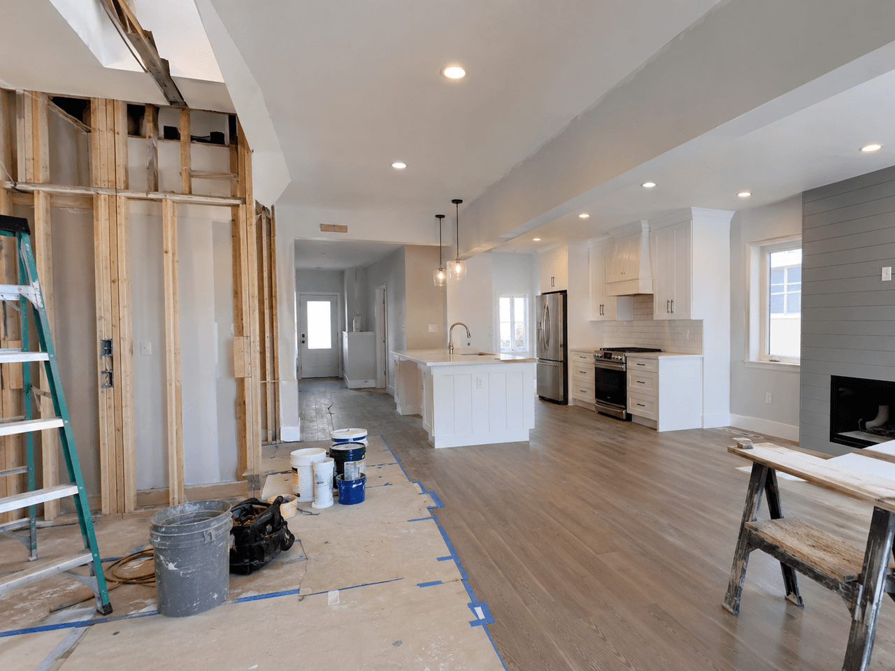 Intérieur à aire ouverte en rénovation avec cuisine blanche, îlot central, plancher en bois, foyer moderne et mur partiellement ouvert laissant voir l’ossature.