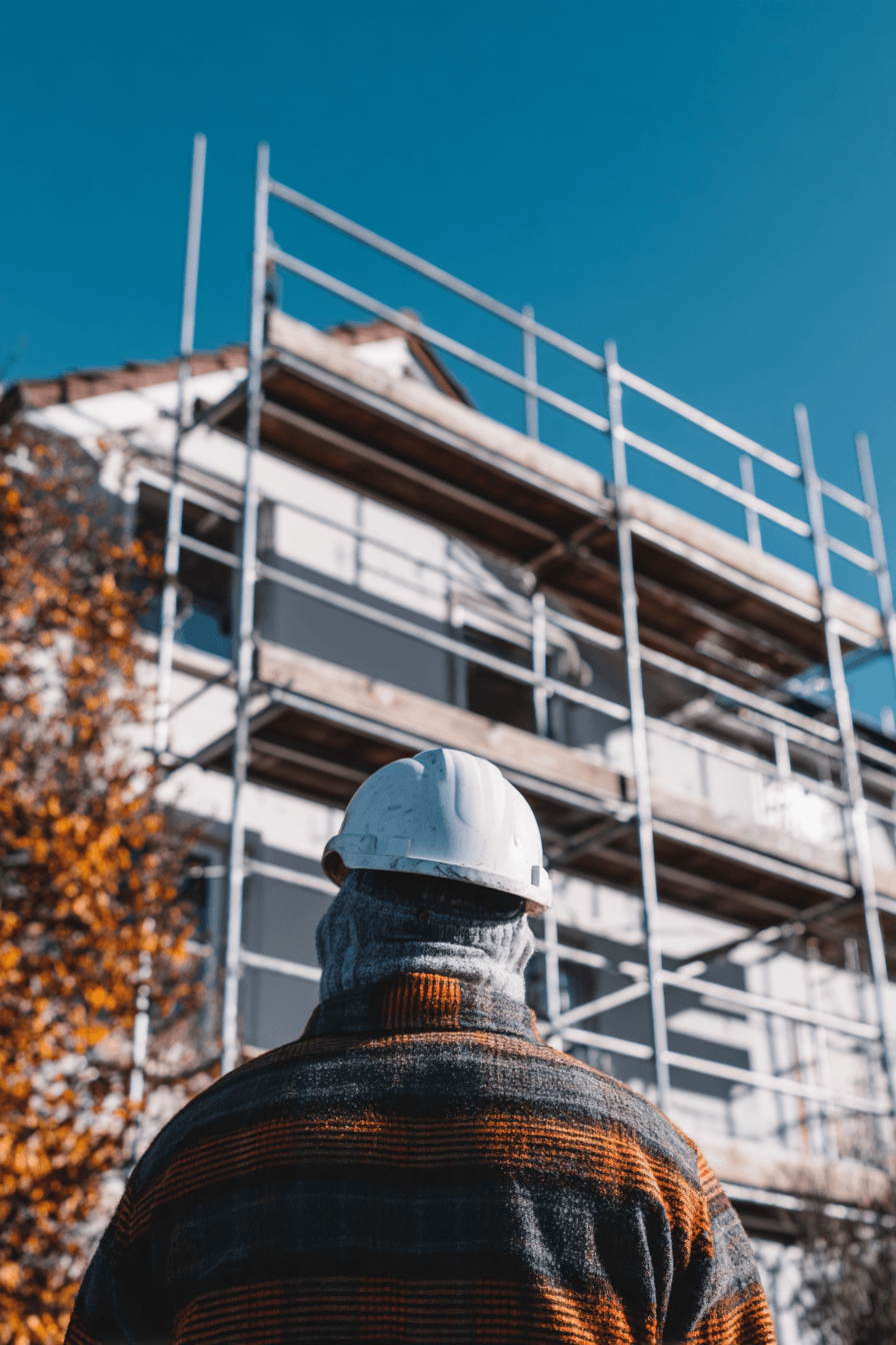 Travailleur de la construction avec casque blanc observant une maison en rénovation extérieure entourée d’échafaudages sous un ciel bleu