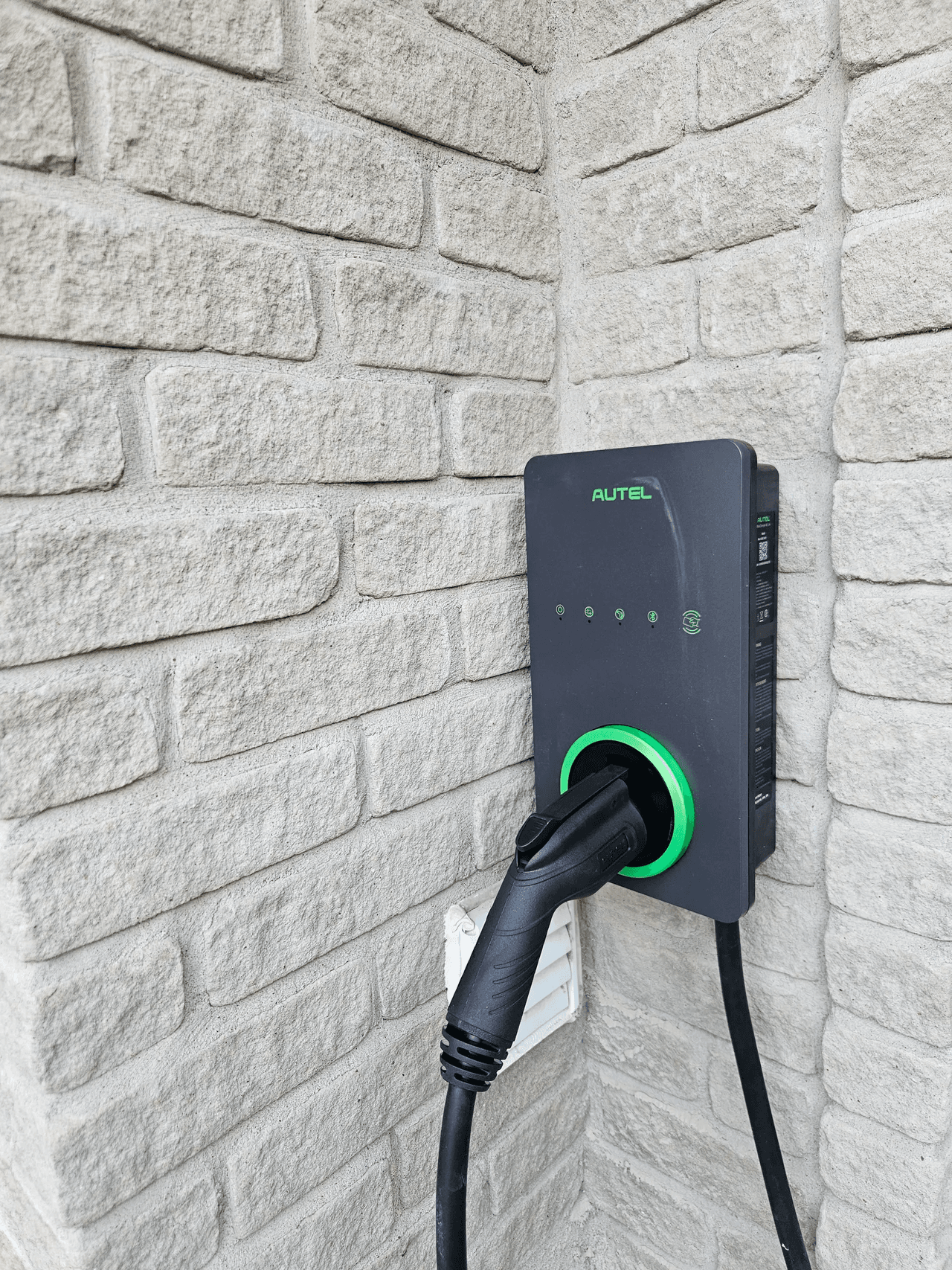 Borne de recharge pour véhicule électrique installée sur un mur extérieur en brique pâle, avec câble de recharge branché.
