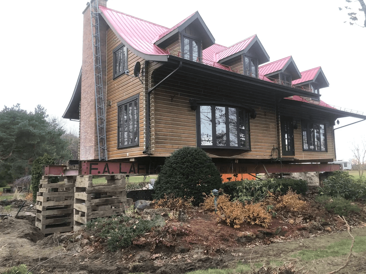Maison en bois surélevée avec toiture métallique rouge, cheminée en brique, fenêtres noires et aménagement paysager en chantier