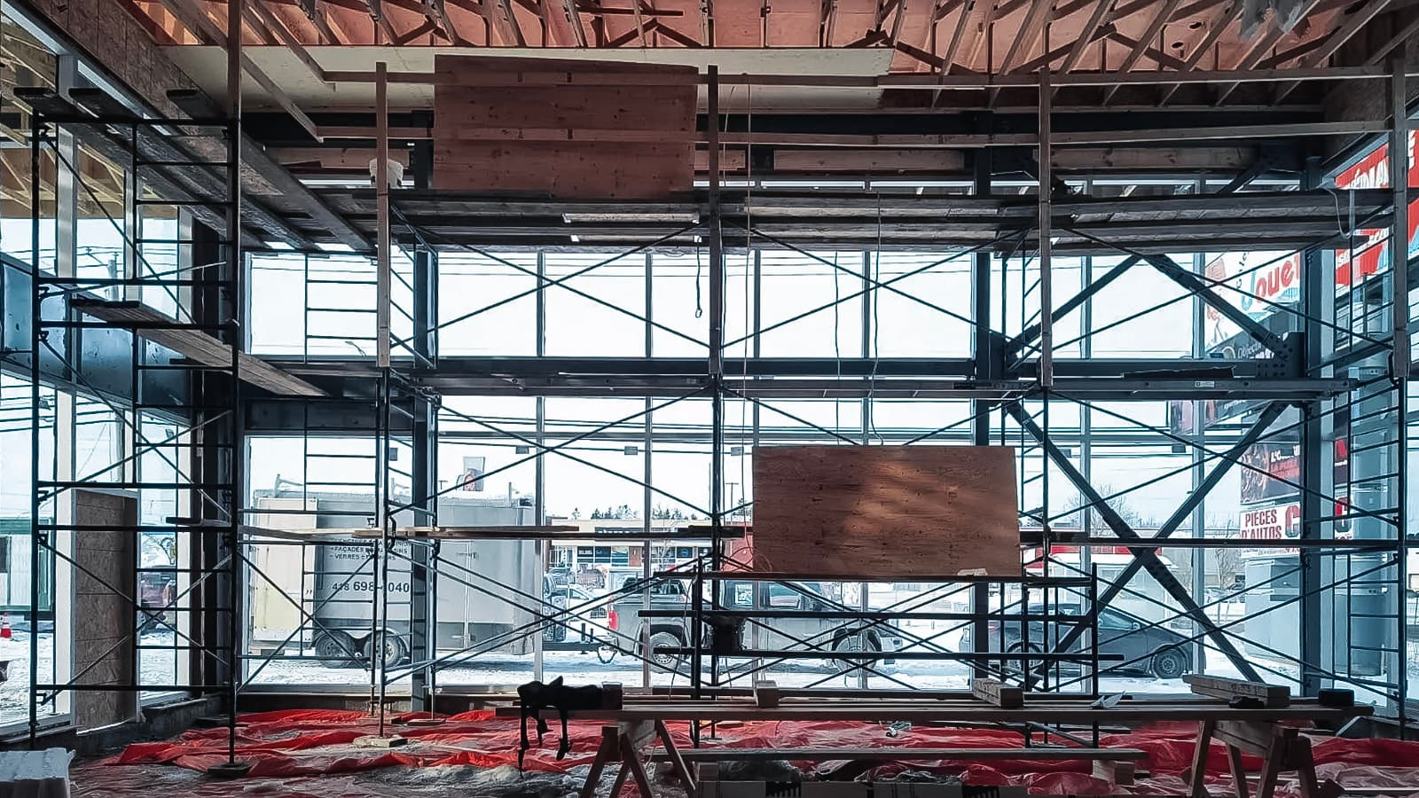 Chantier de construction intérieur avec échafaudages métalliques, grandes fenêtres vitrées, structures en bois et sol recouvert d’une bâche rouge.