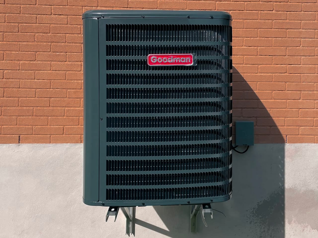 Unité de climatisation extérieure noire de marque Goodman fixée sur un mur de briques oranges avec base en béton.