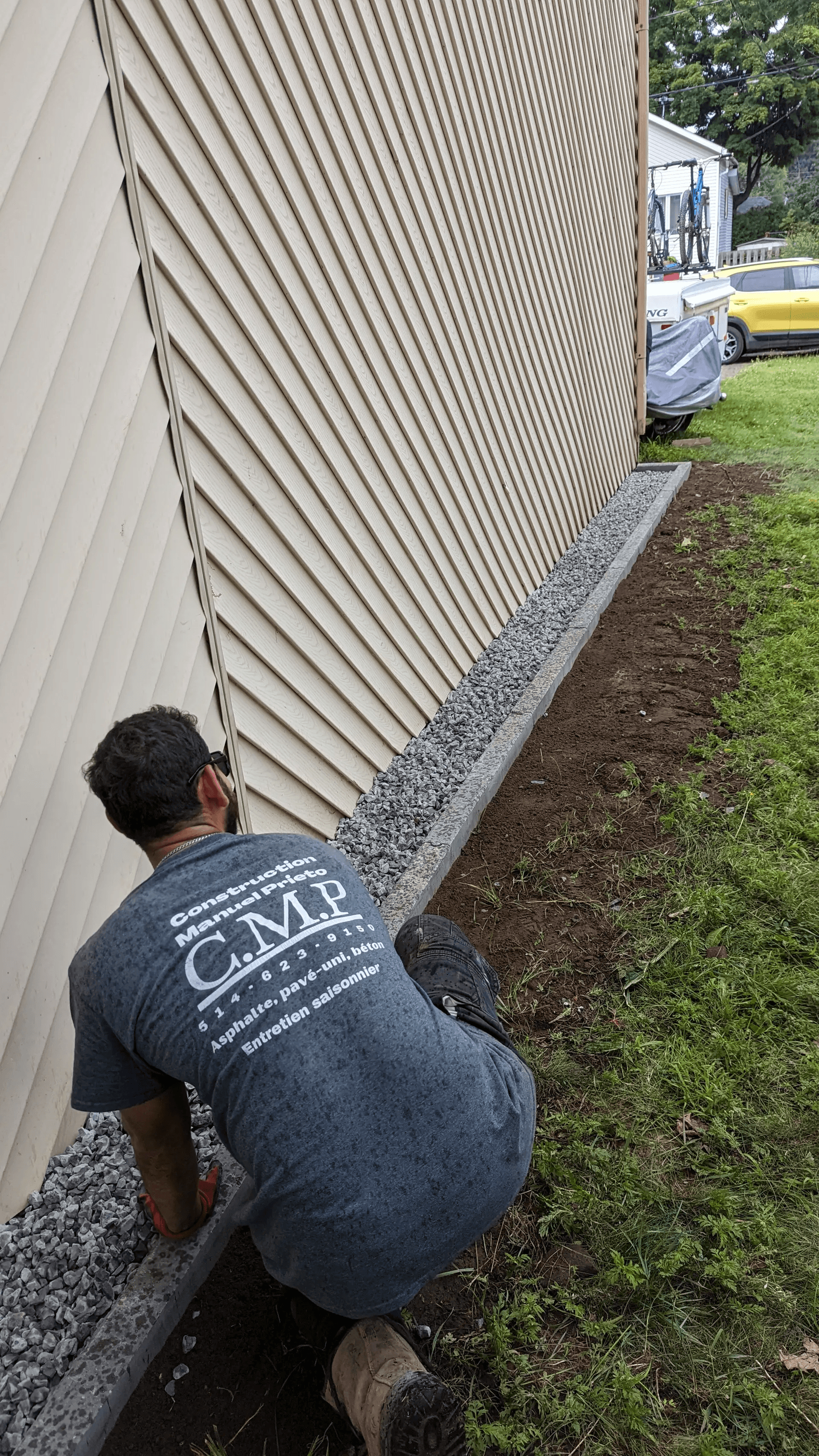 Travaux d’aménagement extérieur résidentiel avec un entrepreneur agenouillé installant une bordure de béton le long du revêtement extérieur en vinyle beige d’une maison, délimitant une bande de pierres concassées grises destinée au drainage et à la finition paysagère des fondations
