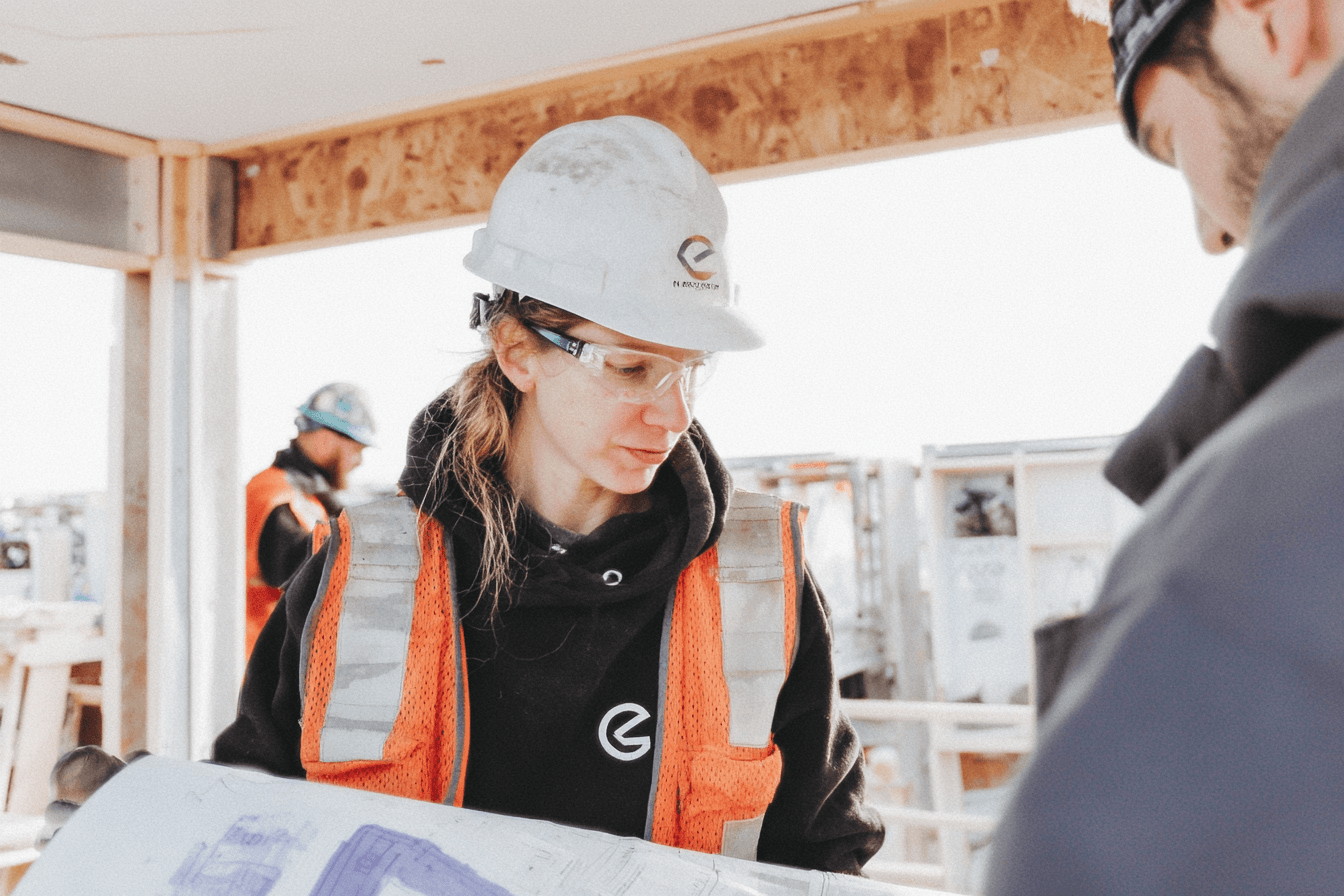 Professionnelle de la construction portant un casque de sécurité, des lunettes de protection et un gilet haute visibilité, consultant des plans sur un chantier