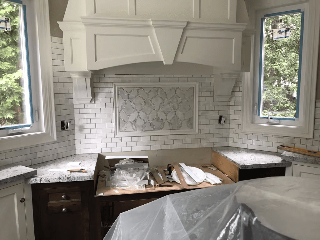 Cuisine en rénovation avec hotte décorative blanche sur mesure, dosseret en carreaux métro, comptoirs de pierre, fenêtres neuves et armoires en cours d’installation.