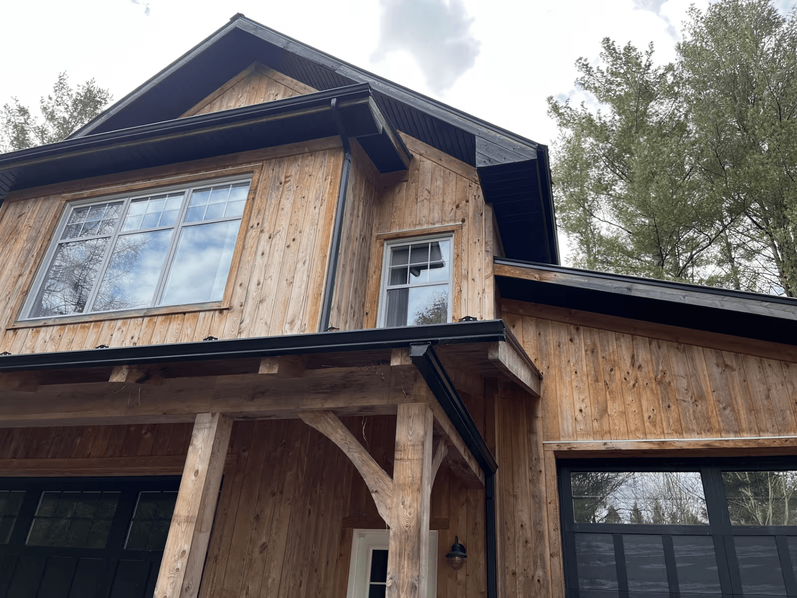 Façade d’une maison résidentielle à deux étages avec revêtement extérieur en bois naturel vertical, toiture en bardeaux foncés, soffites et fascias noirs, gouttières noires, grandes fenêtres blanches, portes de garage noires et porche d’entrée avec poutres et colonnes en bois massif