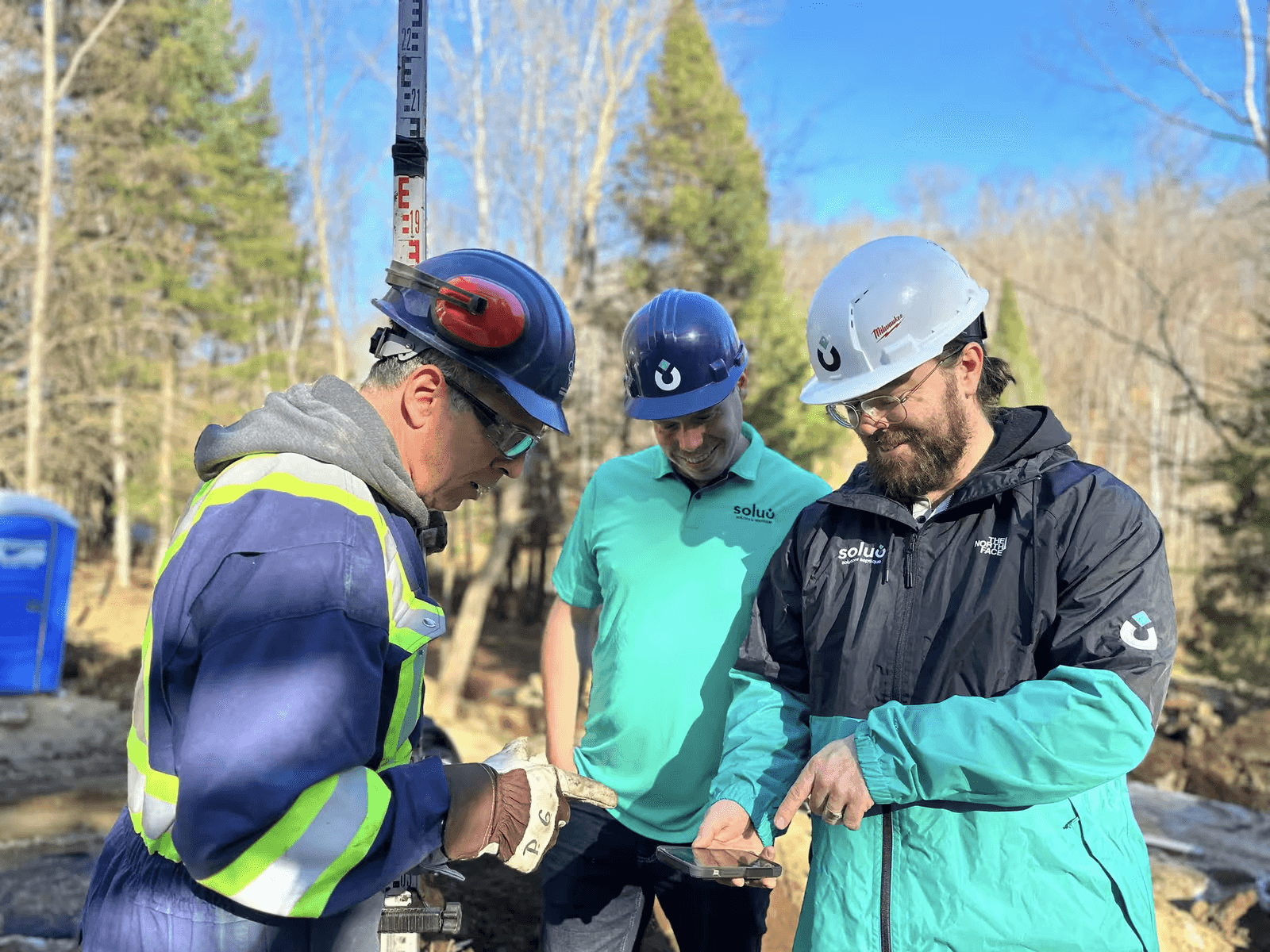 Trois professionnels de chantier portant des casques de sécurité examinent ensemble des informations sur un téléphone mobile dans une zone de travaux extérieure boisée.