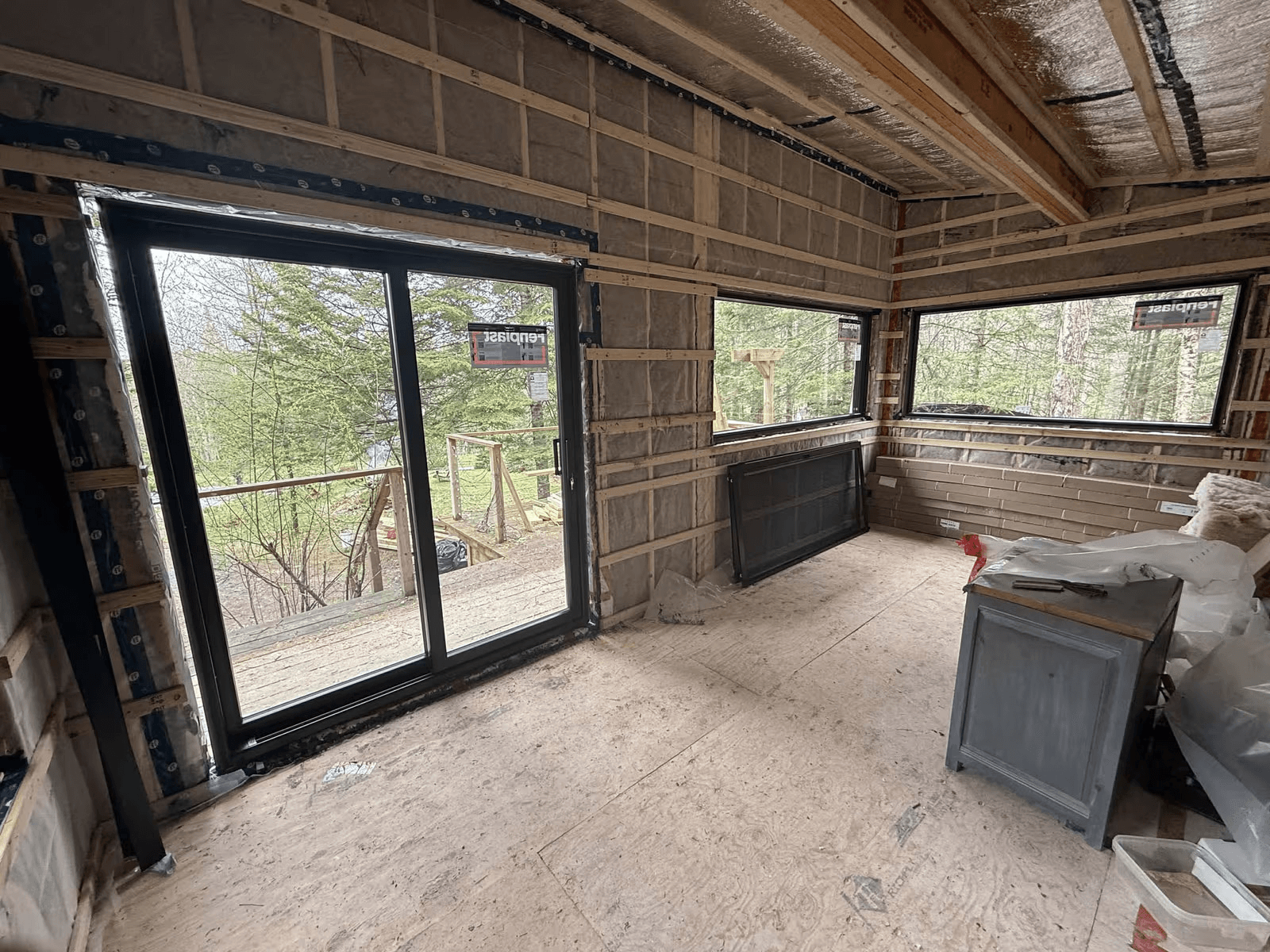 Pièce résidentielle en rénovation avec grande porte-patio noire, fenêtres panoramiques, murs ouverts avec isolation apparente, ossature de bois et vue sur un terrain boisé.