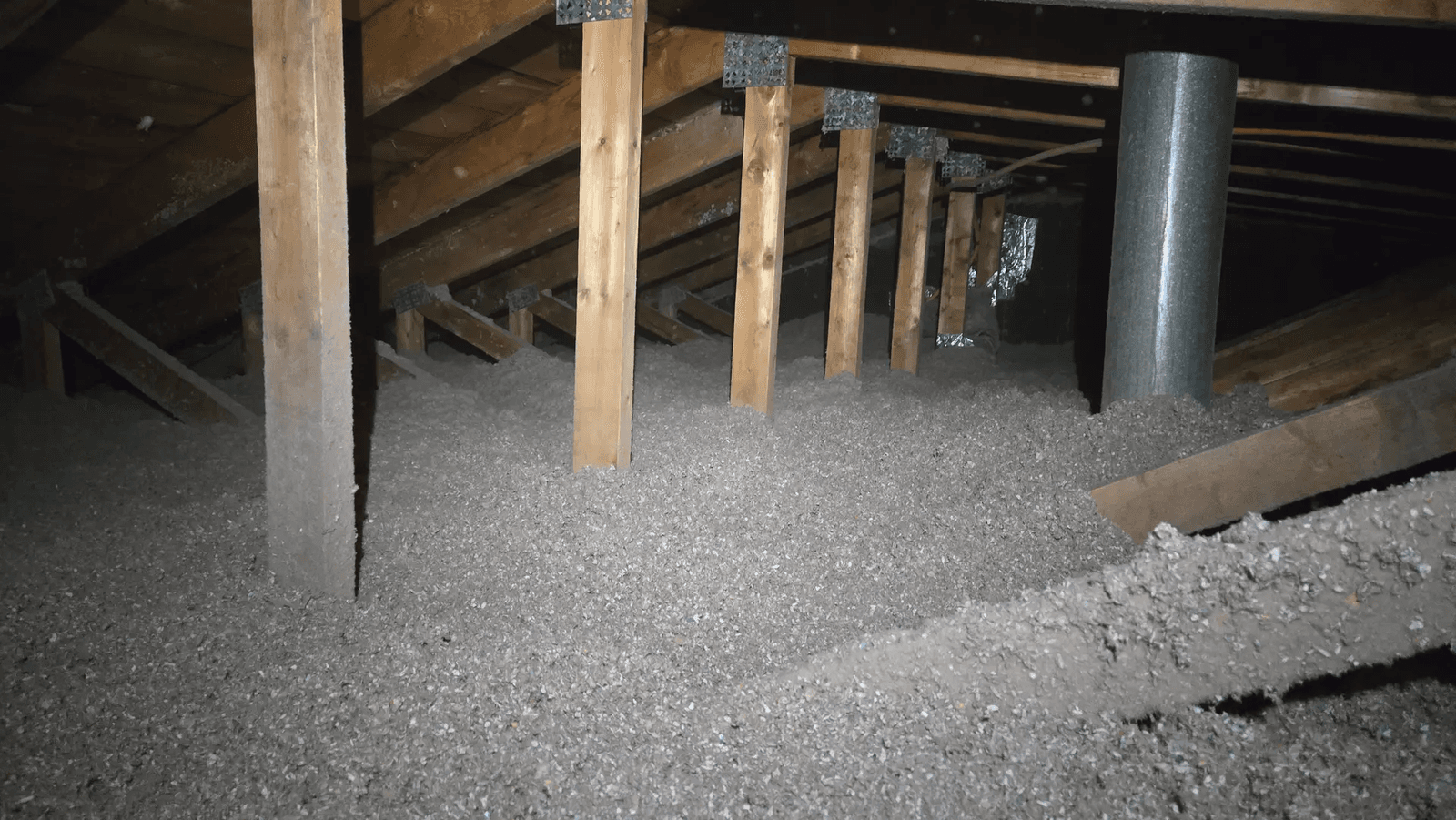 Entretoit résidentiel avec isolation soufflée grise répartie entre les éléments de charpente en bois, montants de soutien, fermes de toit et conduit métallique de ventilation, illustrant des travaux d'isolation thermique des combles pour améliorer l'efficacité énergétique d'une maison