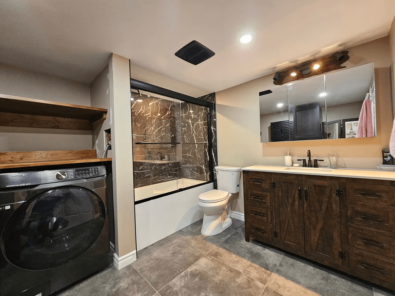 Salle de bain rénovée avec douche vitrée en céramique effet marbre noir, vanité en bois foncé, lavabo double, plancher en tuiles grises et espace buanderie intégré