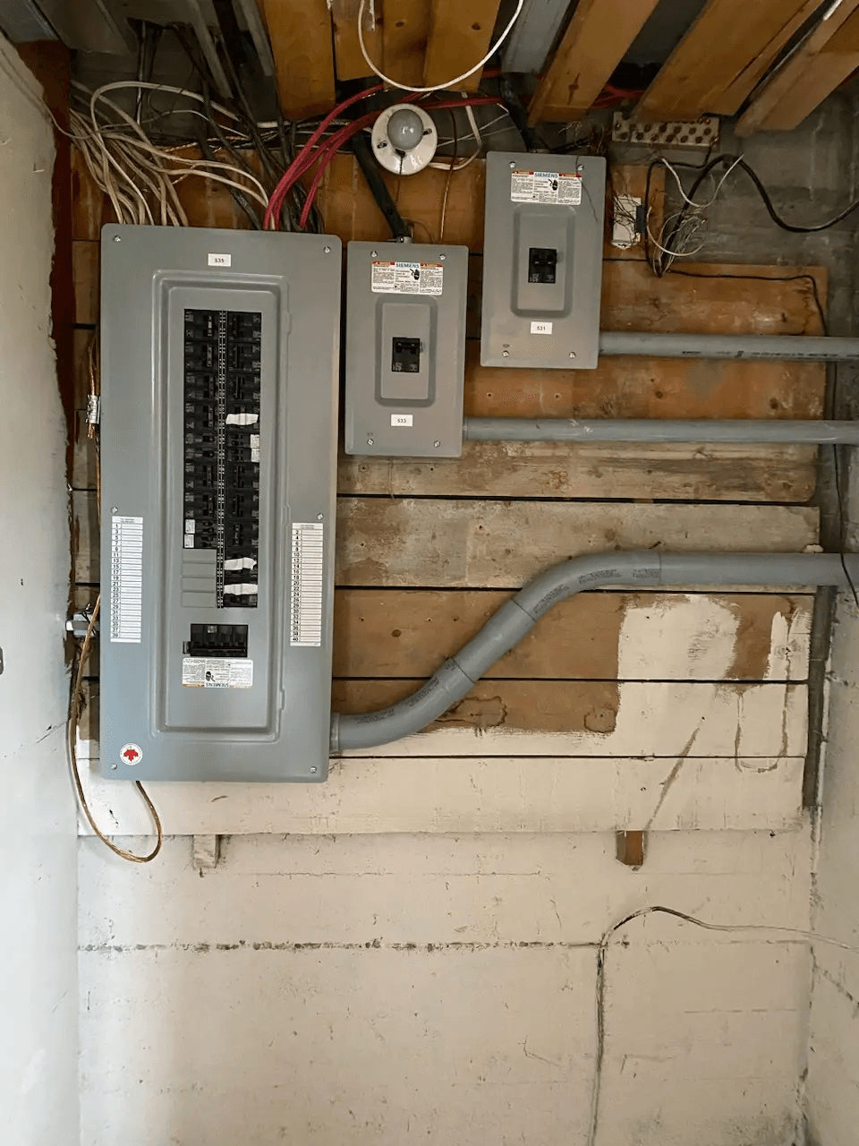 Panneau électrique principal et sous-panneaux installés sur un mur en bois brut avec conduits gris et câblage apparent