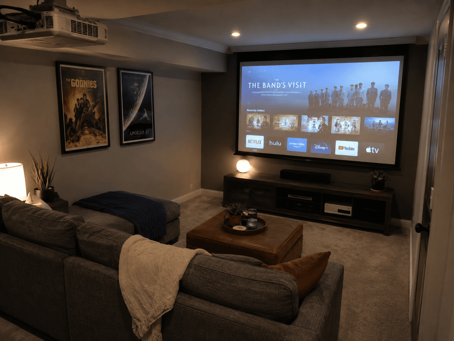 Salle de cinéma maison avec projecteur au plafond, grand écran de projection, canapé d'angle, meuble multimédia et affiches de films encadrées.