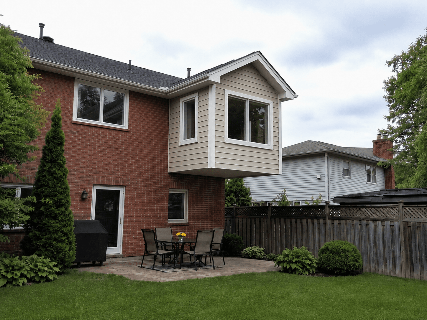Agrandissement arrière en porte-à-faux d’une maison en brique avec grandes fenêtres, revêtement beige, toiture à pignon et cour aménagée avec patio