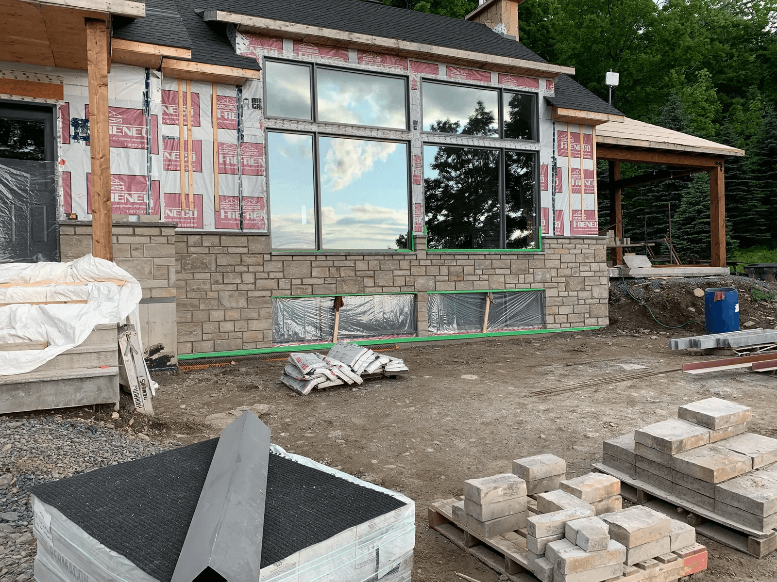 Maison en construction avec revêtement de pierre partiellement installé, grandes fenêtres et matériaux de chantier au sol