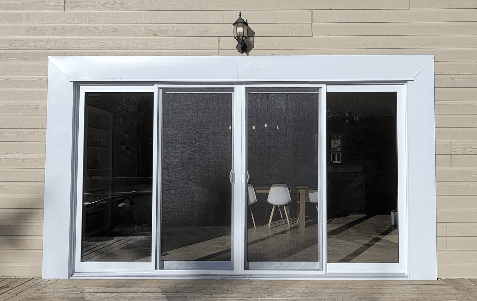 Grande porte-patio coulissante en verre avec cadre blanc, moustiquaires centrales et revêtement extérieur horizontal beige donnant sur une terrasse en bois.