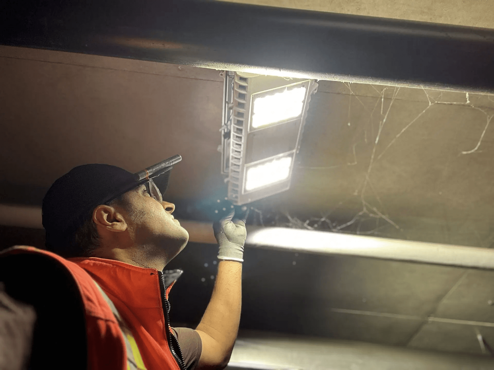 Électricien inspectant ou réparant un luminaire LED au plafond dans un espace sombre avec lampe frontale et gants de sécurité.