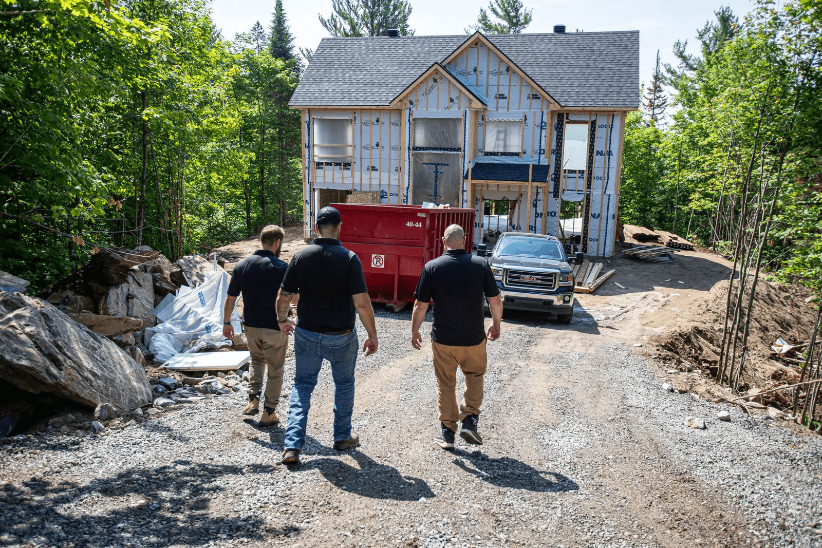 Trois professionnels inspectant un chantier de construction résidentielle avec une maison en construction entourée de végétation.