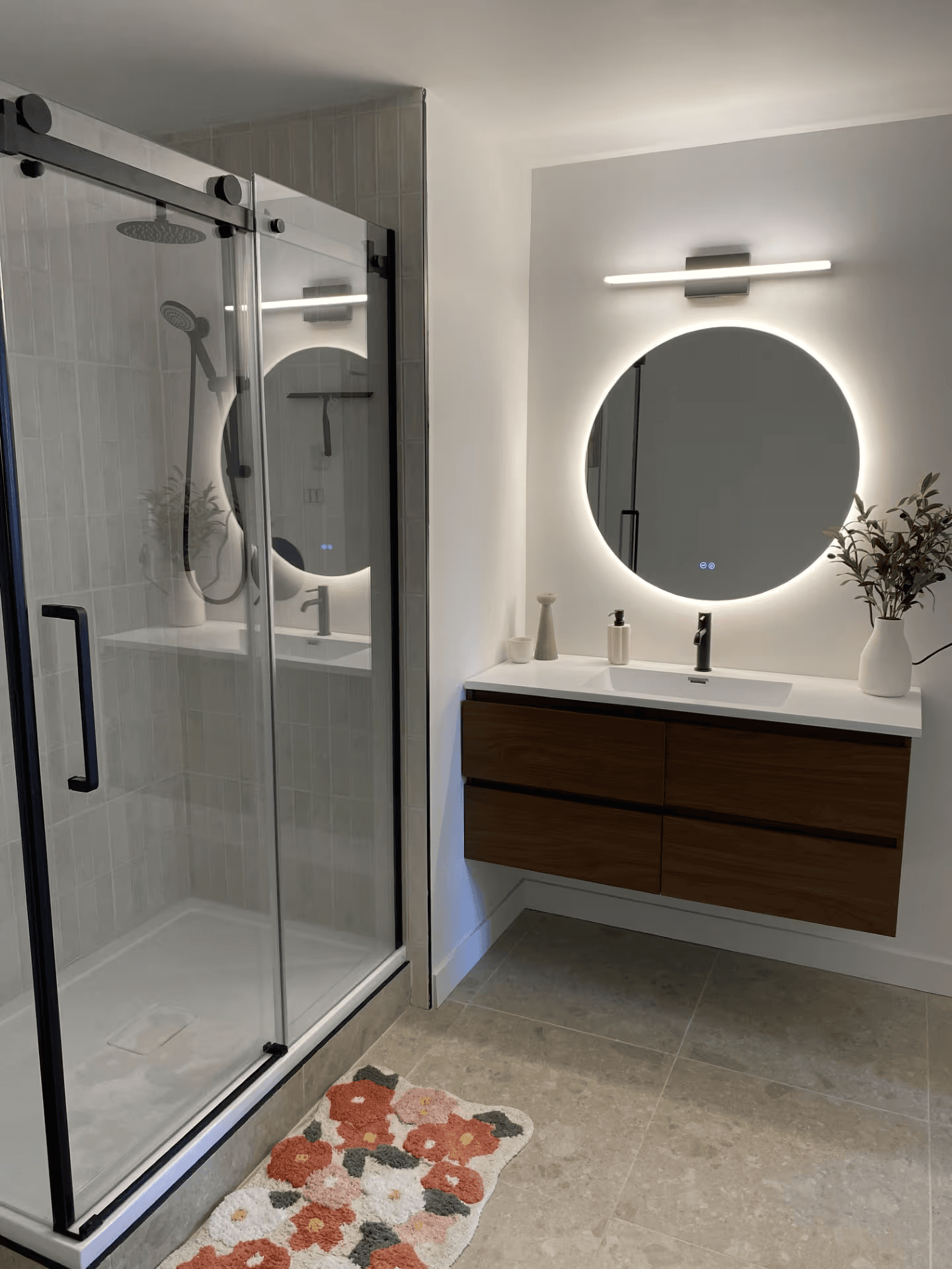 Salle de bain moderne rénovée avec douche vitrée, robinetterie noire, vanité suspendue en bois et miroir rond lumineux