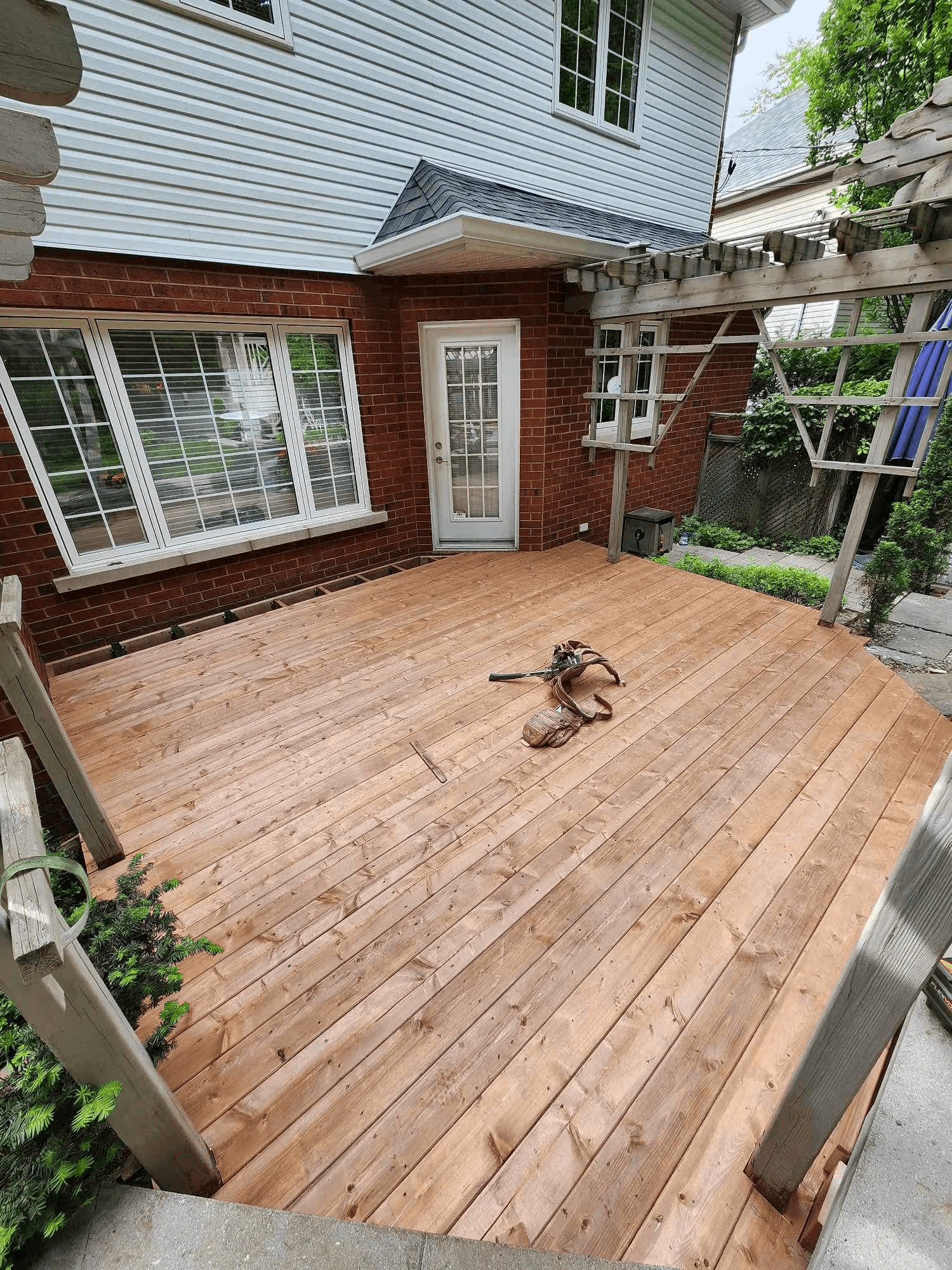 Grande terrasse en bois récemment construite à l’arrière d’une maison en brique et revêtement blanc, avec pergola existante et accès par porte vitrée