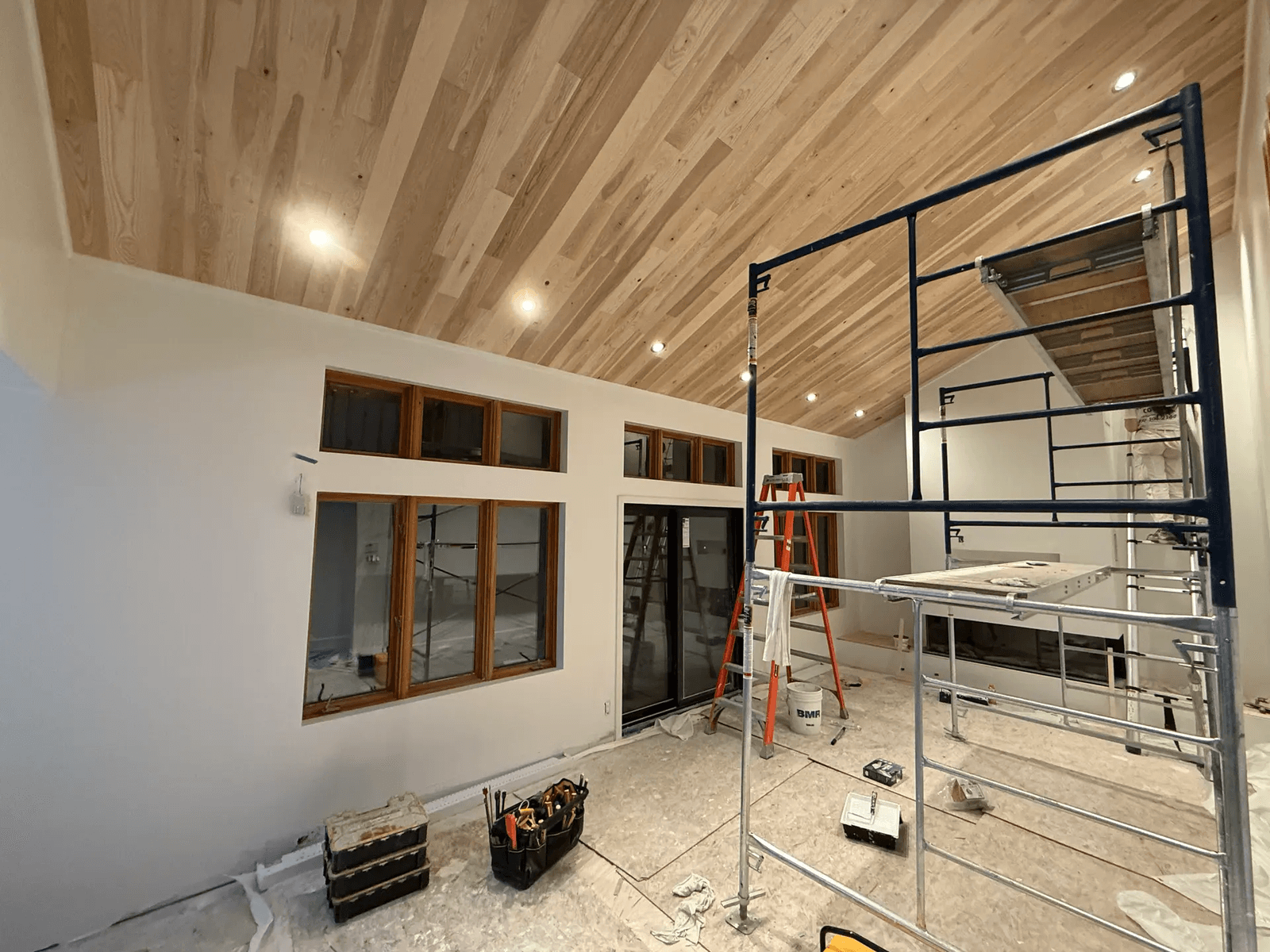 Pièce résidentielle en rénovation avec plafond incliné en bois, éclairage encastré, grandes fenêtres et échafaudage installé pour les travaux de finition