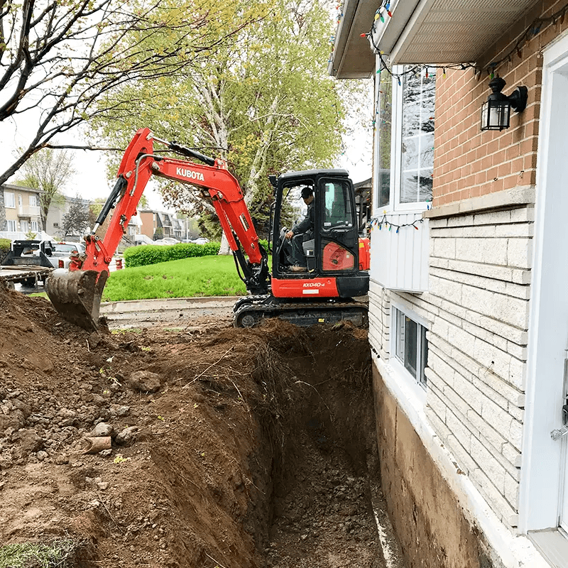 Travaux d’excavation résidentielle avec une mini-excavatrice Kubota orange creusant une profonde tranchée le long des fondations en béton d’une maison en brique, illustrant la préparation du terrain pour l’installation ou la réparation d’un drain français et l’imperméabilisation des fondations