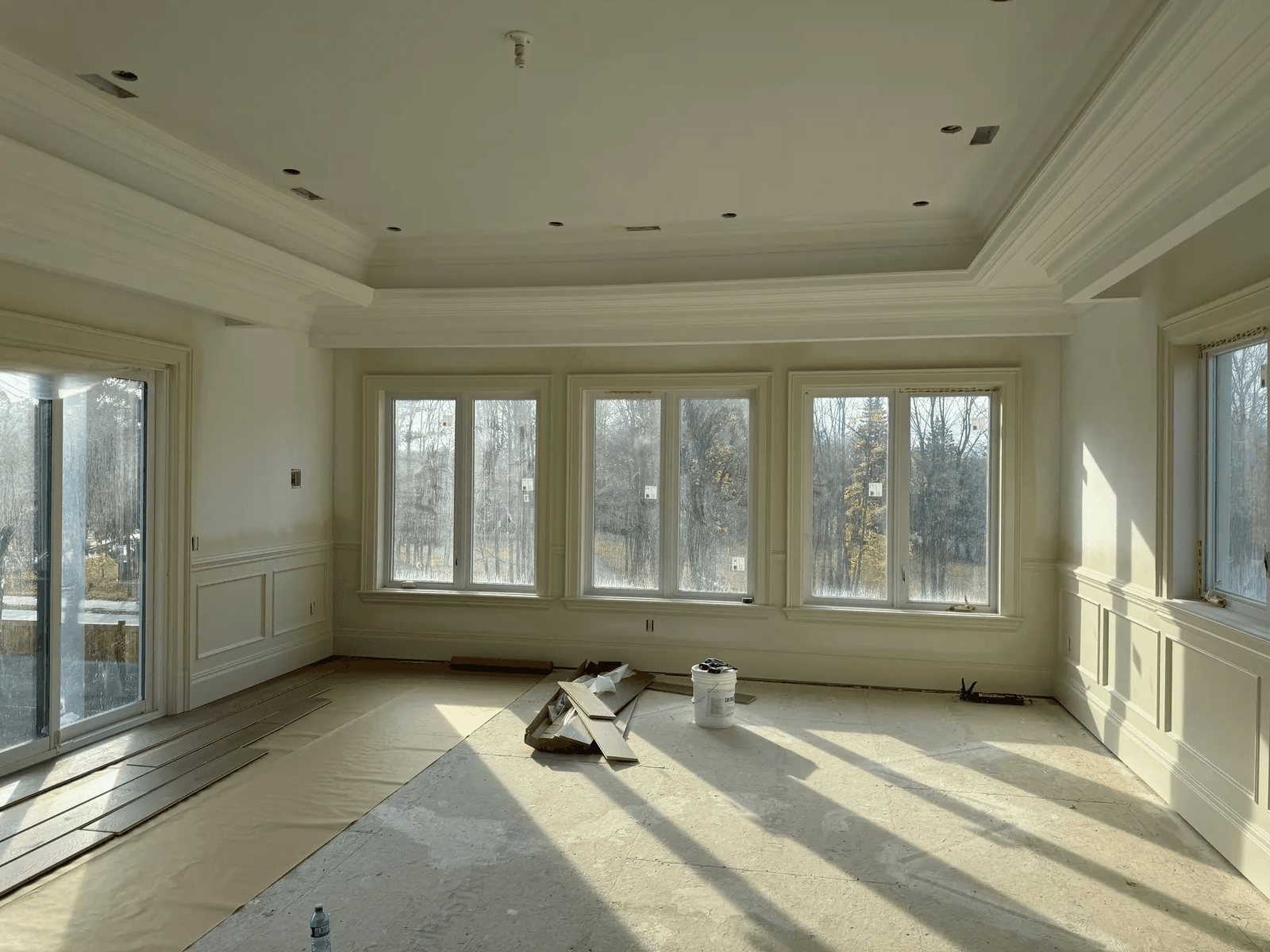 Pièce lumineuse en rénovation avec moulures décoratives, grandes fenêtres, plafond à caissons et plancher en cours d’installation.