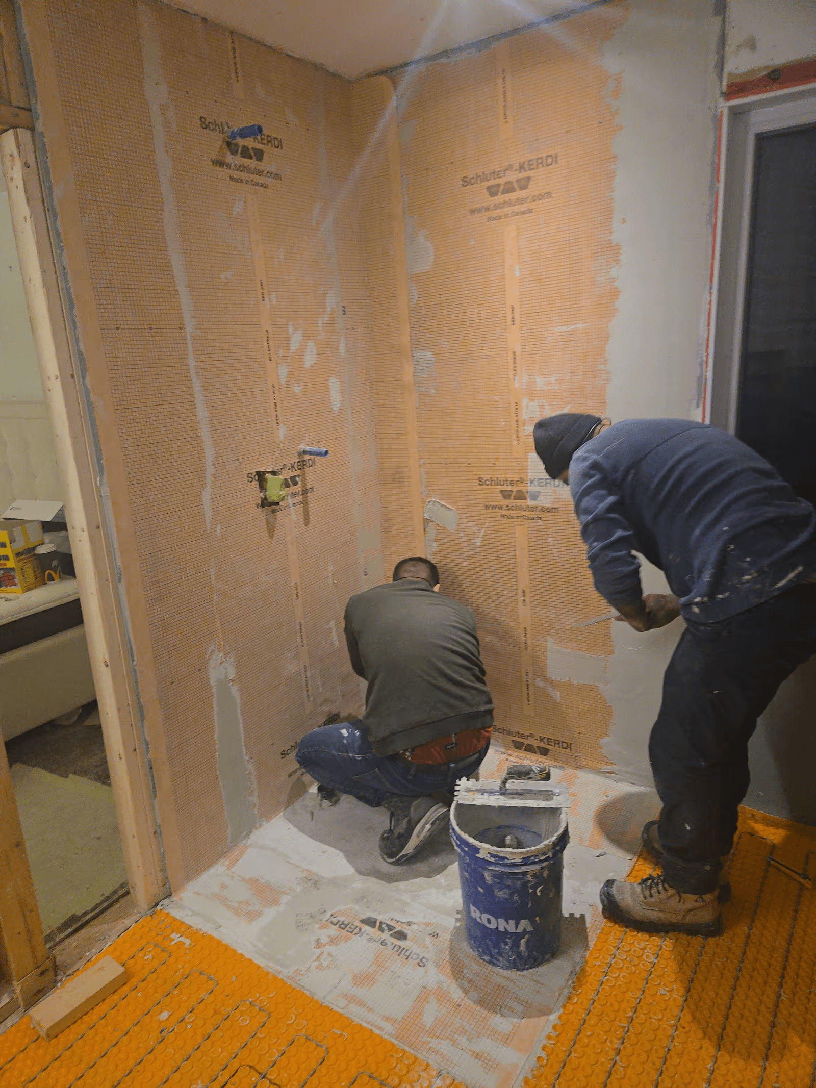 Installation d’une membrane d’étanchéité orange dans une douche en rénovation par deux ouvriers, avec préparation des murs et du plancher avant la pose de céramique