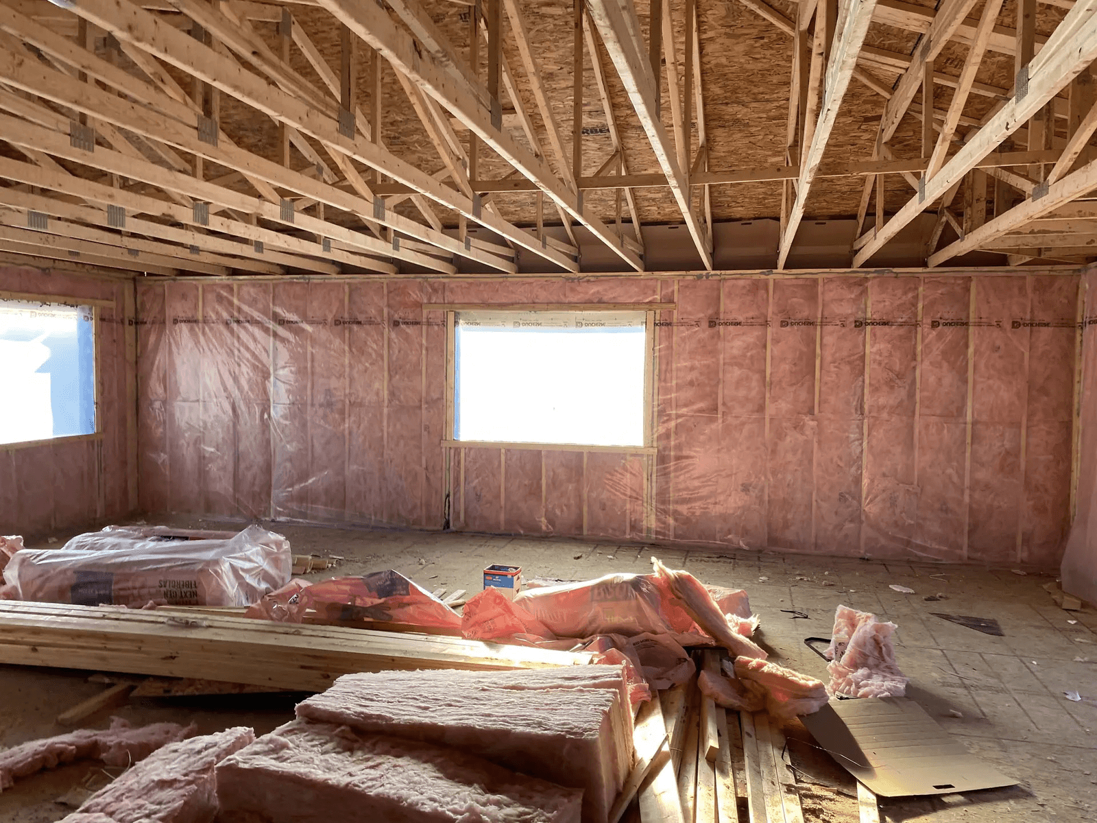 Intérieur résidentiel en construction avec charpente de toiture apparente, murs isolés en fibre de verre rose, pare-vapeur et matériaux de chantier au sol.
