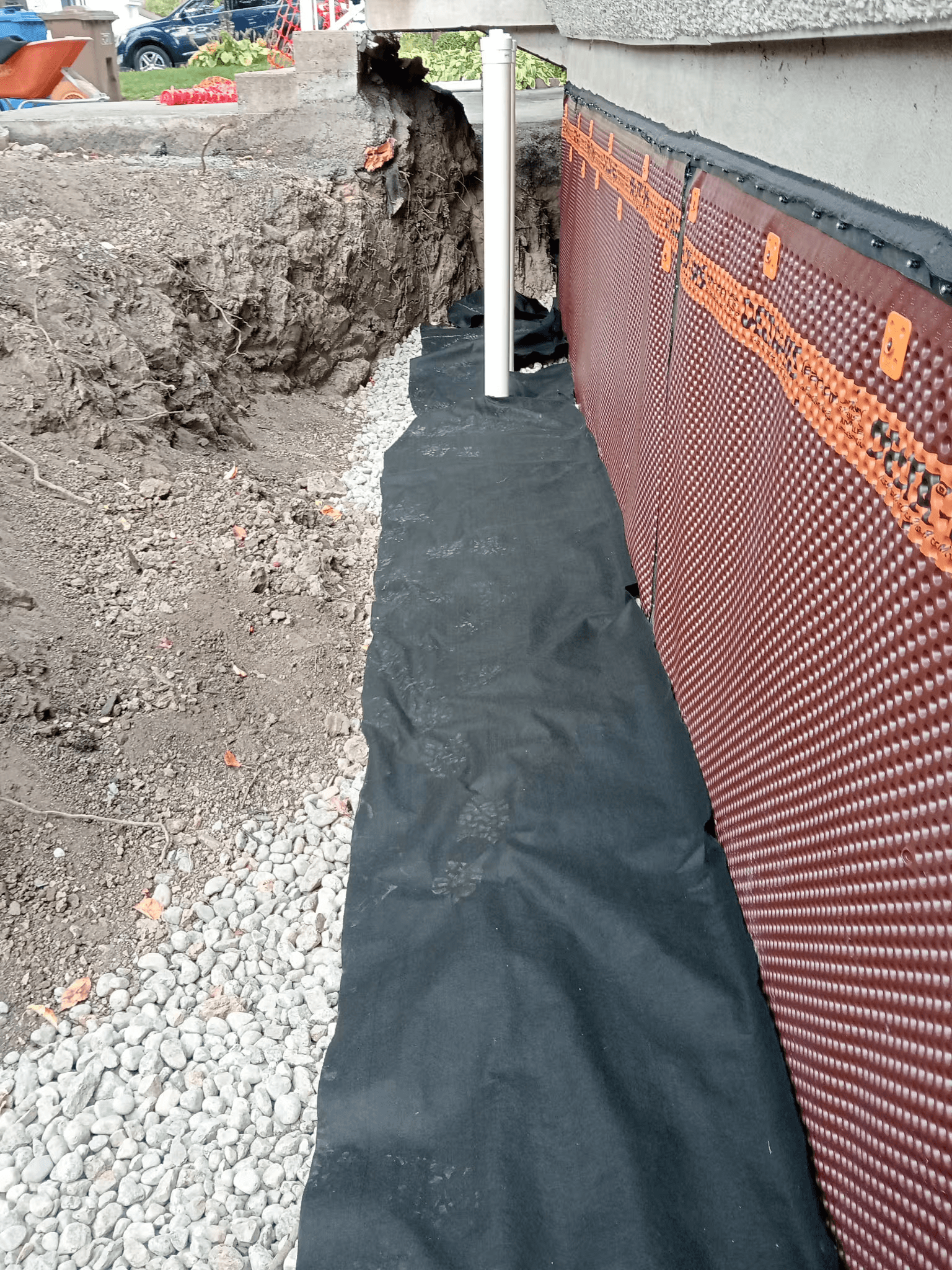 Travaux d’imperméabilisation des fondations d’une maison résidentielle avec installation d’une membrane drainante alvéolée brune sur un mur de béton, membrane géotextile noire recouvrant un lit de pierre concassée et tranchée d’excavation, illustrant la protection des fondations contre l’humidité et les infiltrations d’eau