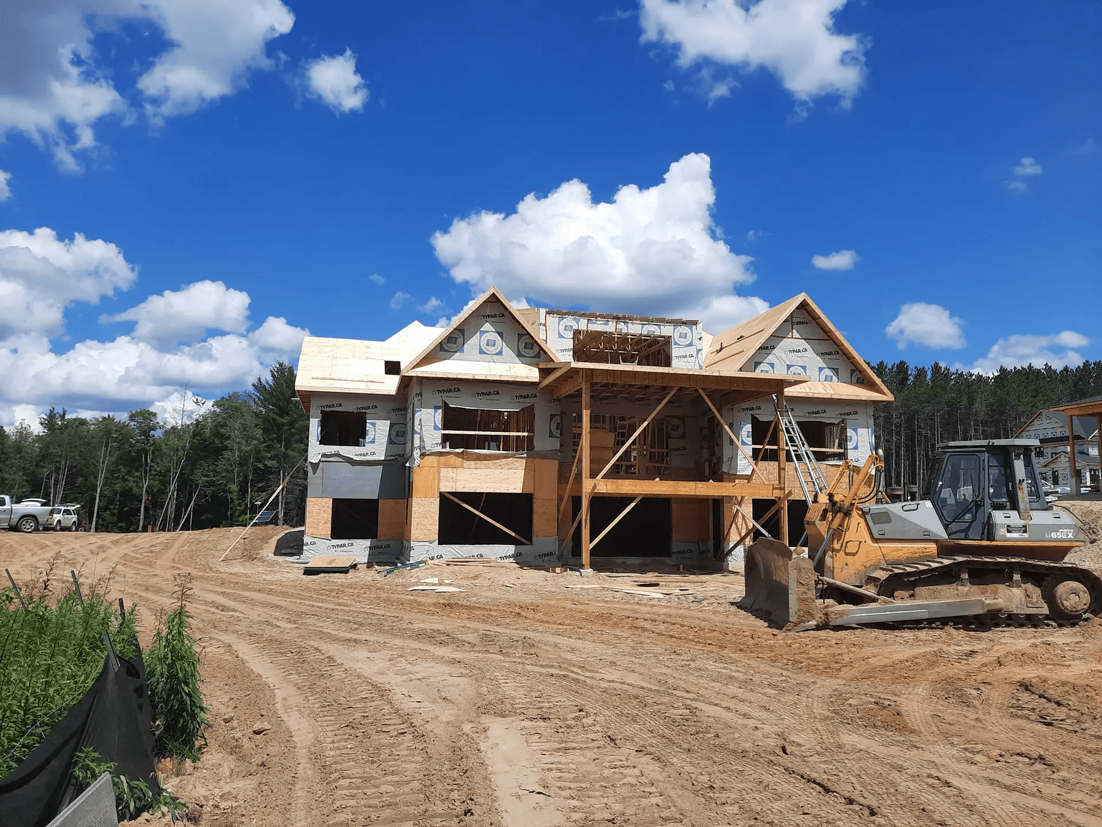 Maison à deux étages en construction sur terrain sablonneux avec pare-intempéries Tyvek, échafaudages en bois et machinerie lourde sur place.