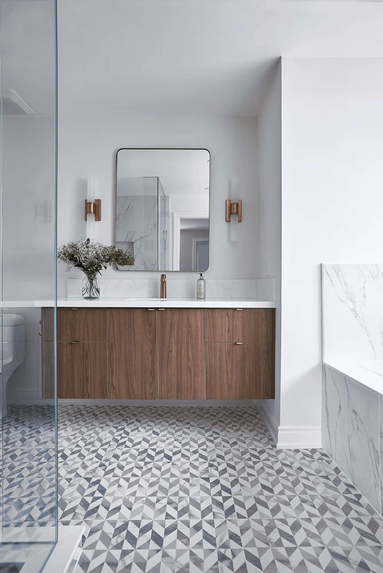 Salle de bain contemporaine haut de gamme avec vanité en bois brun, comptoir blanc, robinetterie dorée, miroir rectangulaire aux coins arrondis, appliques murales dorées, plancher en céramique géométrique gris et blanc, douche vitrée et baignoire habillée de carreaux effet marbre blanc