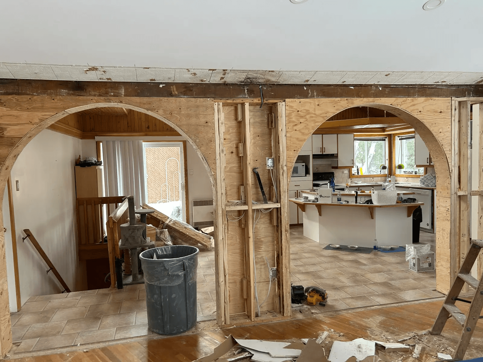 Rénovation intérieure résidentielle en cours avec mur porteur ouvert, grandes arches en bois, structure apparente, câblage électrique et cuisine visible en arrière-plan