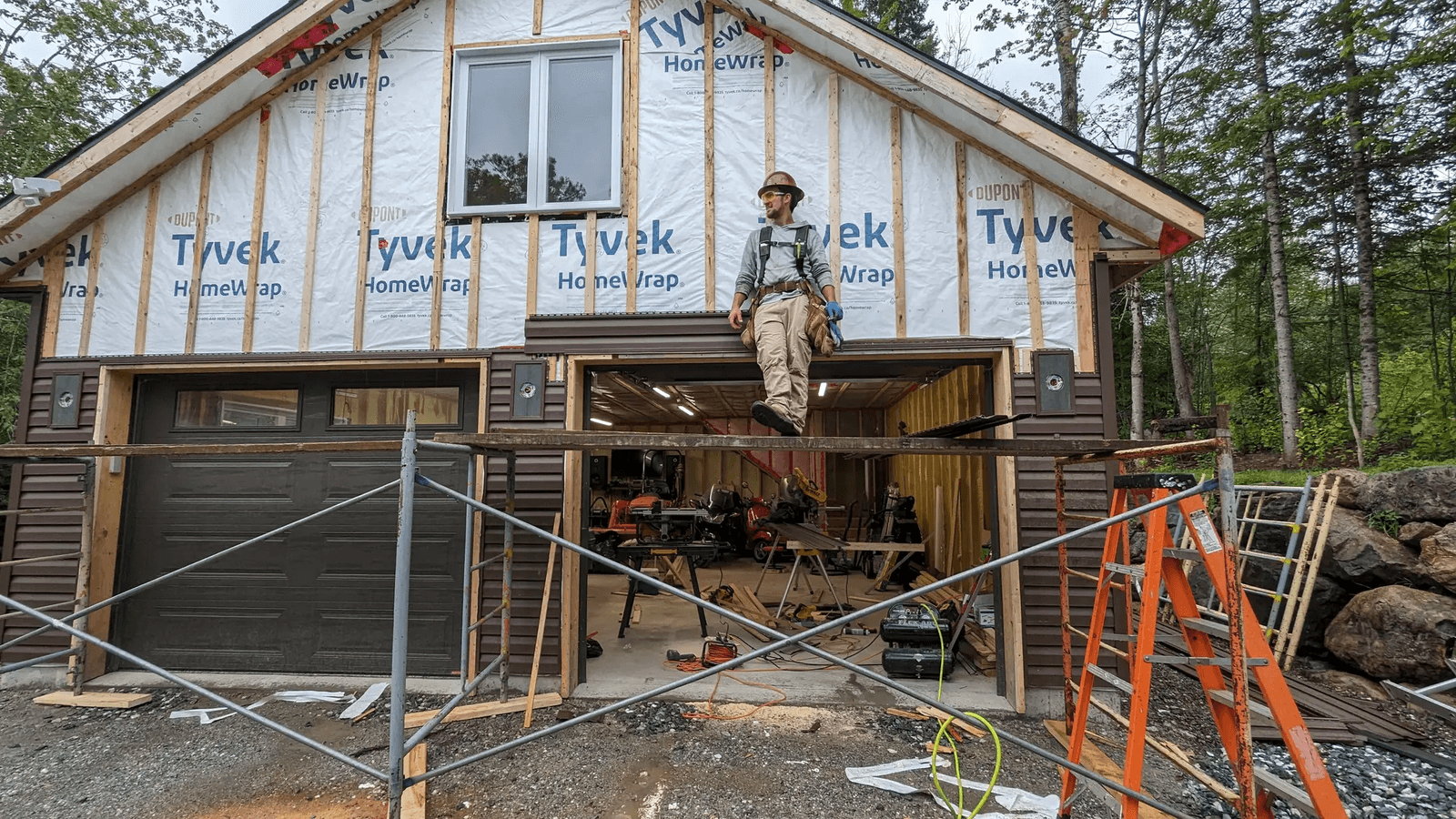 Travailleur de la construction devant une maison en rénovation avec membrane pare-intempéries, échafaudage, échelles, outils et revêtement extérieur partiellement installé