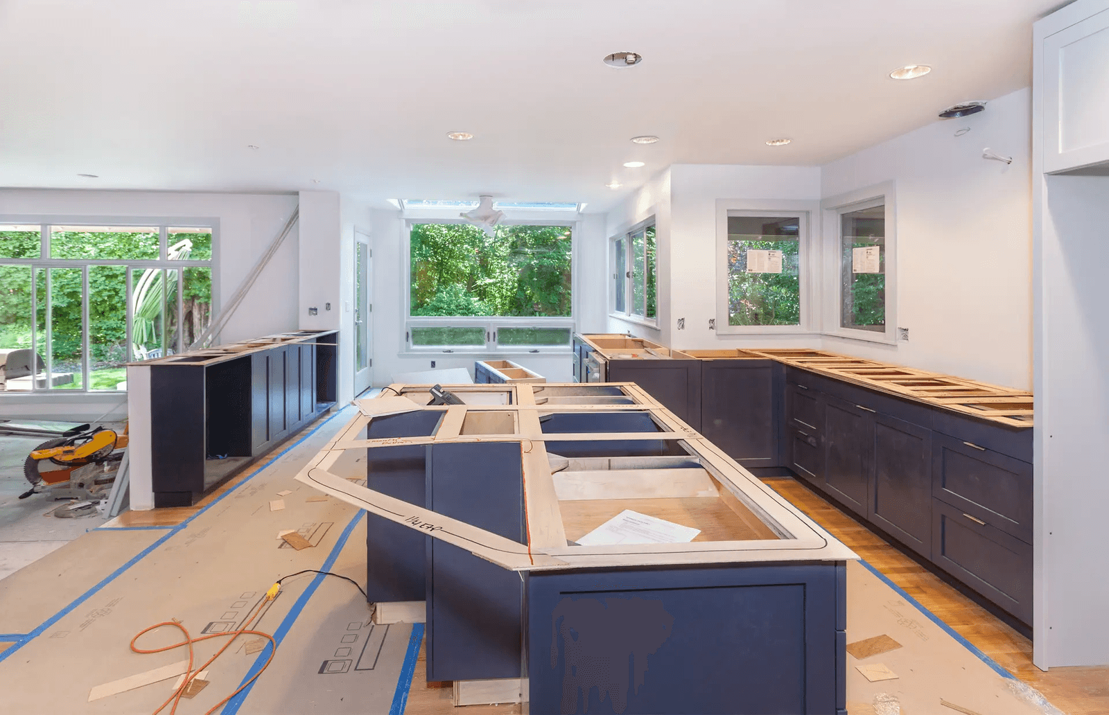 Cuisine résidentielle en rénovation avec armoires bleu foncé en cours d’installation, grand îlot central, planchers protégés, éclairage encastré et grandes fenêtres donnant sur la cour