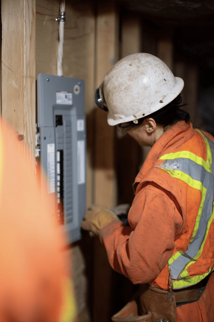 Électricien inspectant un panneau lectrique dans un sous-sol avec gilet de scurit, lampe frontale et outils de travail.