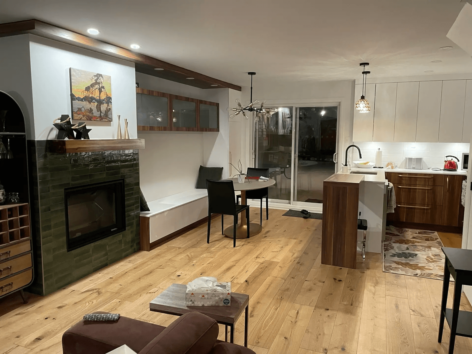 Aire de vie ouverte rénovée avec cuisine moderne, foyer encastré, plancher de bois, coin repas et grande porte vitrée donnant sur l’extérieur.