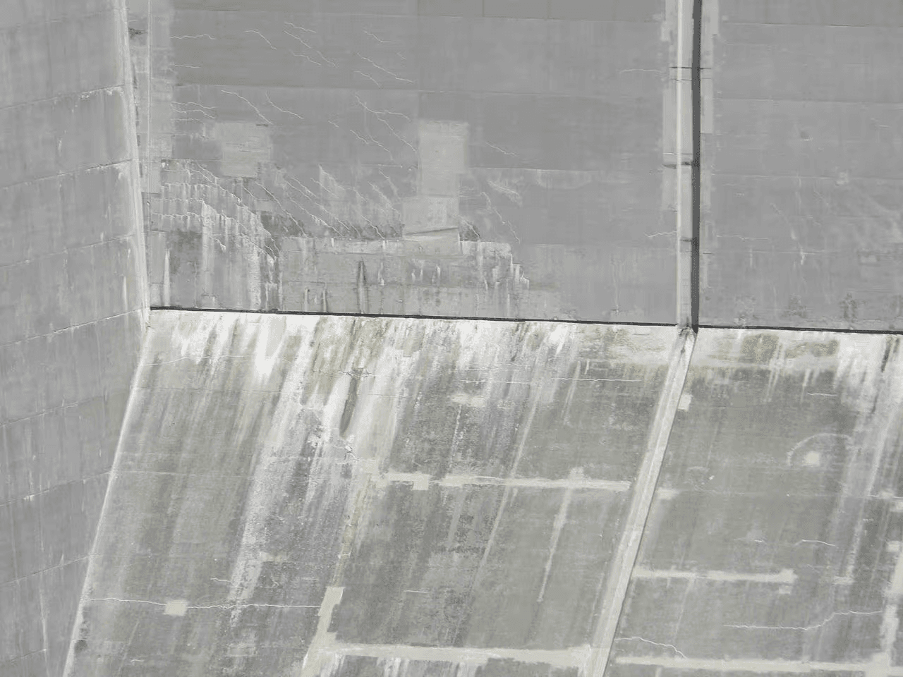 Mur de fondation en béton présentant de fines fissures, des taches d’humidité, des traces de ruissellement et des dépôts minéraux sur une surface en béton inclinée. 