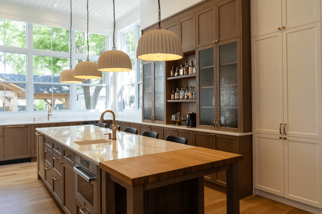 Cuisine haut de gamme avec grand îlot central, armoires en bois et blanches, comptoir clair, luminaires suspendus et nombreuses fenêtres