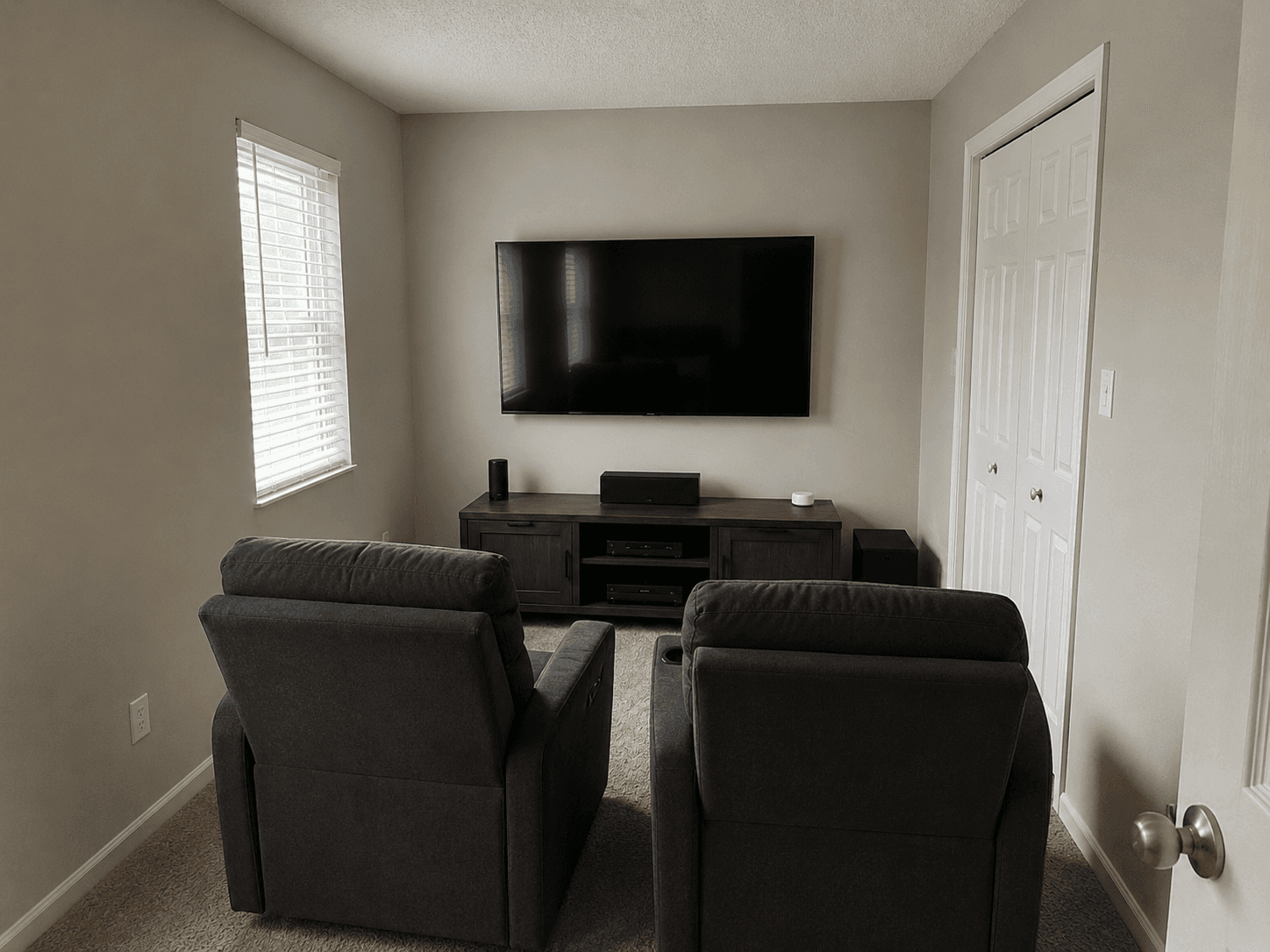 Petit home cinéma aménagé dans une pièce résidentielle, comprenant une télévision murale, un meuble multimédia, un système audio, deux fauteuils inclinables et de la moquette.