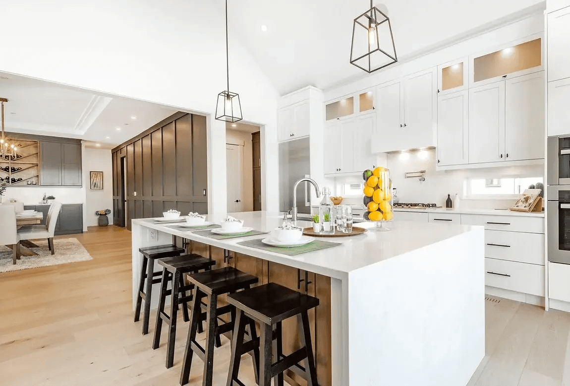 Cuisine moderne blanche avec grand îlot central, tabourets noirs, armoires sur mesure, luminaires suspendus et plancher de bois clair