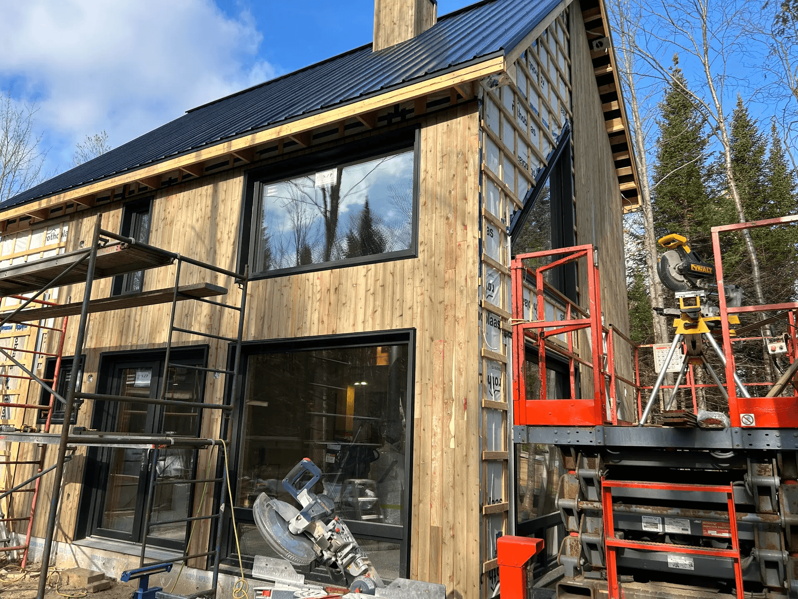 Maison en construction avec revêtement extérieur en bois, grandes fenêtres et toit en tôle noire, entourée d’échafaudages et d’outils.