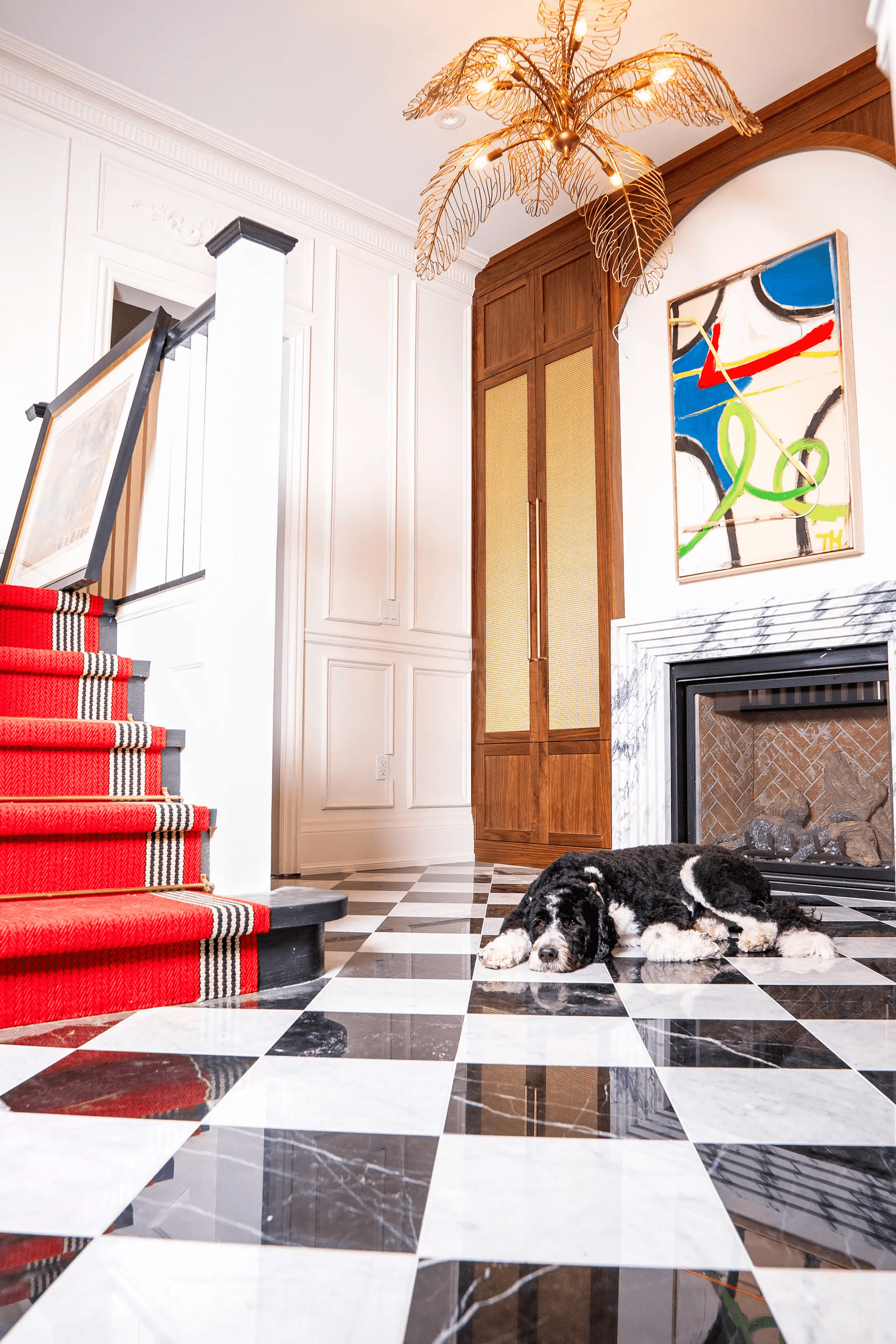 Hall d’entrée élégant avec escalier tapissé de rouge, foyer en marbre, lustre doré et sol en damier noir et blanc