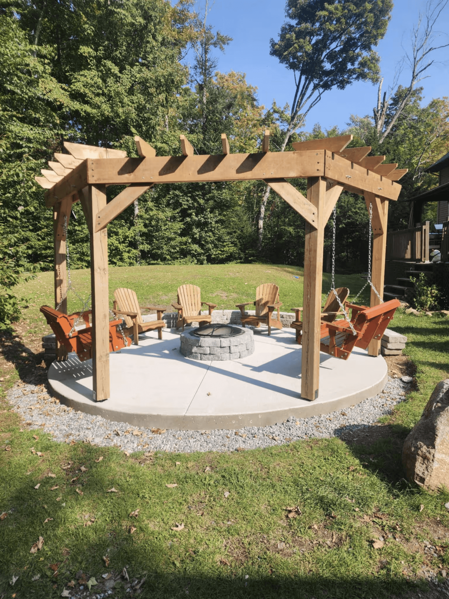 Pergola en bois aménagée sur dalle de béton circulaire avec foyer en pierre, chaises Adirondack suspendues et espace détente dans une cour boisée.
