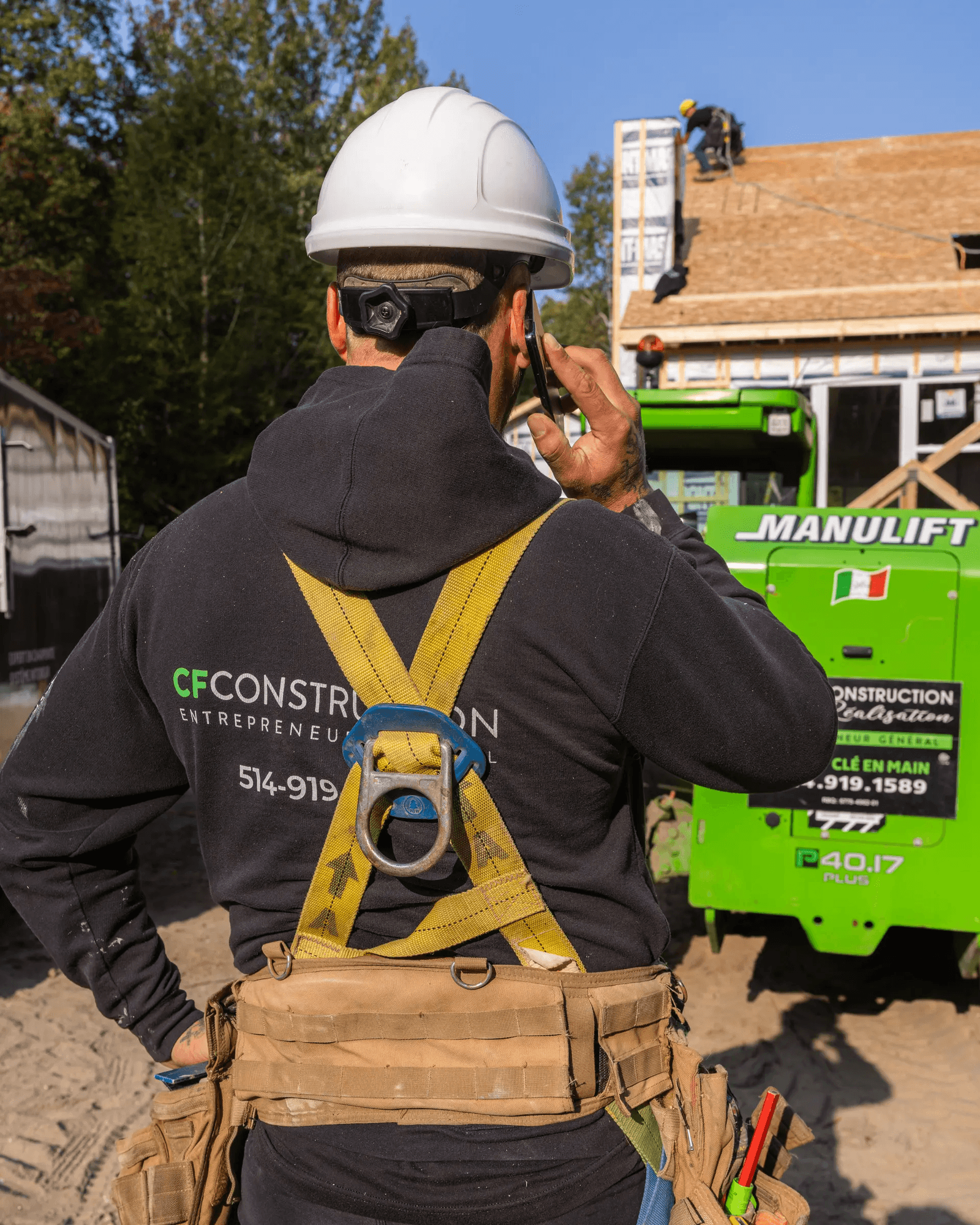Travailleur de la construction portant un harnais de sécurité et un casque, en communication téléphonique sur un chantier avec des travaux de toiture en arrière-plan.