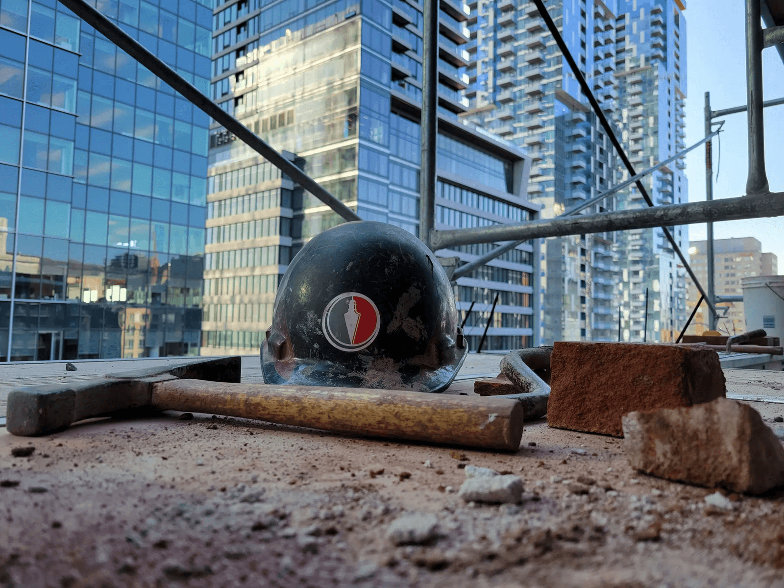  Vue d'un chantier en hauteur avec un casque de sécurité, un marteau et des briques, entouré d'immeubles modernes en arrière-plan.