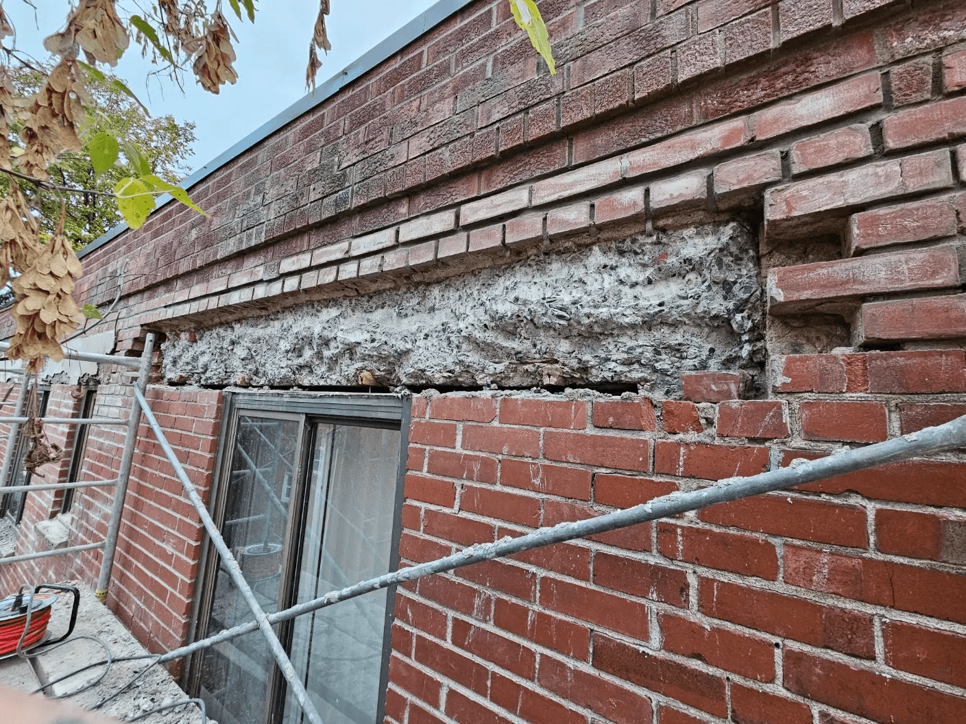 Mur extérieur en brique en cours de réparation avec maçonnerie retirée au-dessus d’une fenêtre, béton exposé et échafaudage installé le long du bâtiment