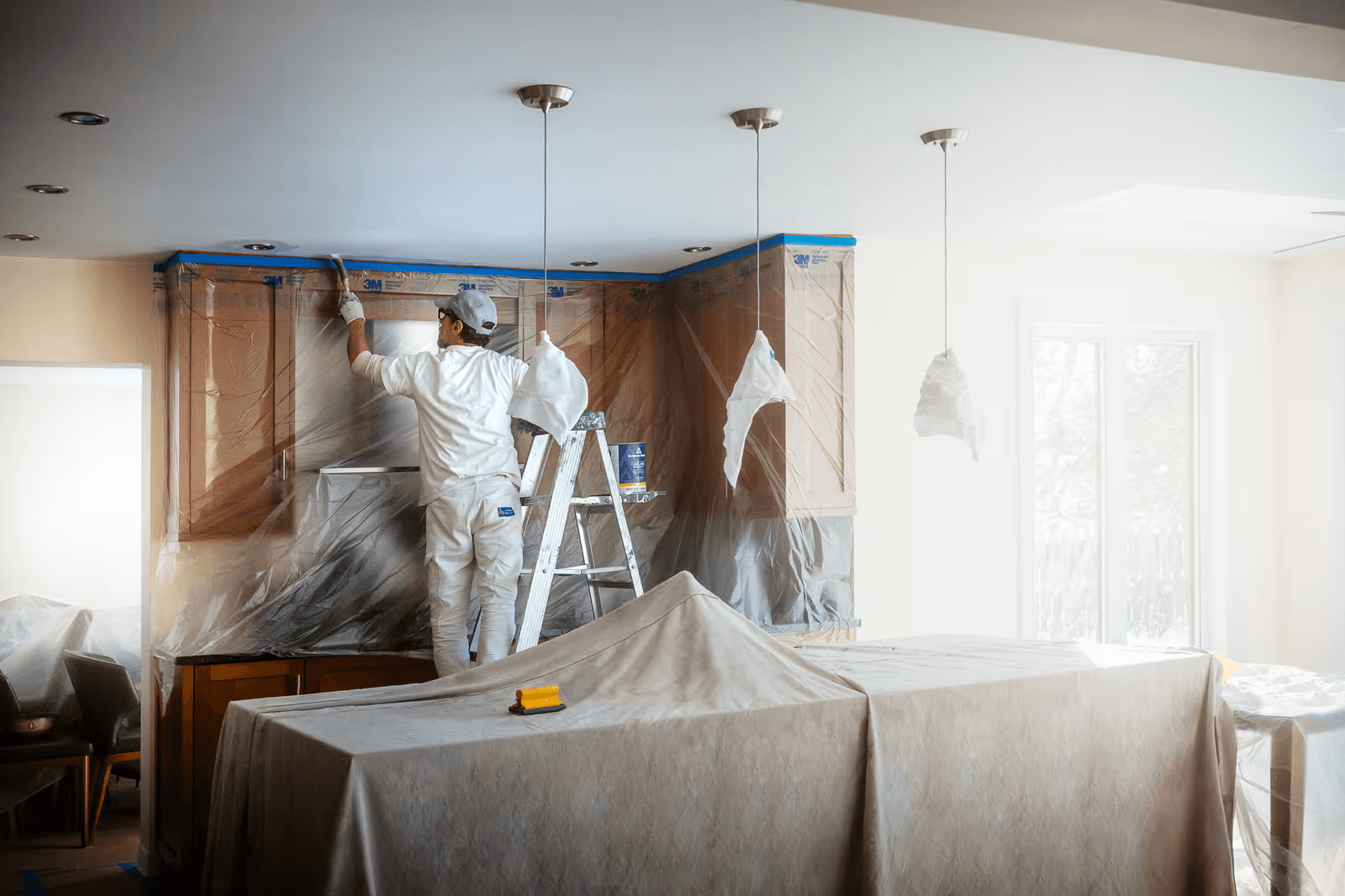 Peintre professionnel préparant et peignant une cuisine résidentielle avec armoires protégées par des bâches et surfaces couvertes pendant les travaux