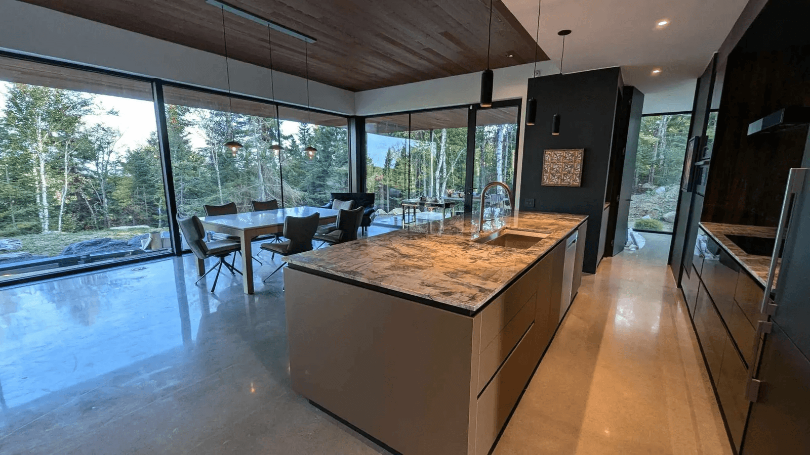 Cuisine moderne avec îlot central en marbre, grandes baies vitrées offrant une vue sur la forêt, plafond en bois et sol en béton poli.