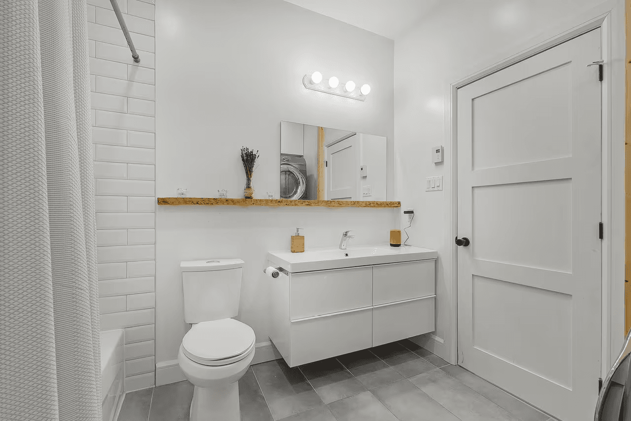 Salle de bain moderne et épurée avec meuble-lavabo blanc suspendu, miroir mural, toilette, douche carrelée, plancher gris et porte intérieure blanche 